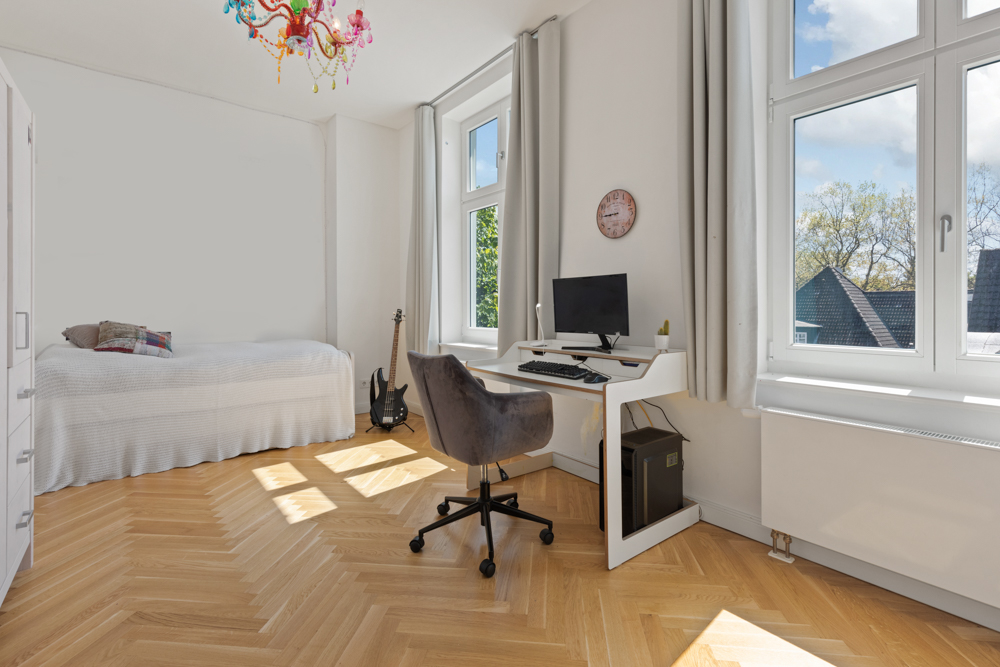 Helles Zimmer mit Einzelbett, Schreibtisch, Computer, Stuhl und Gitarre am Fenster.