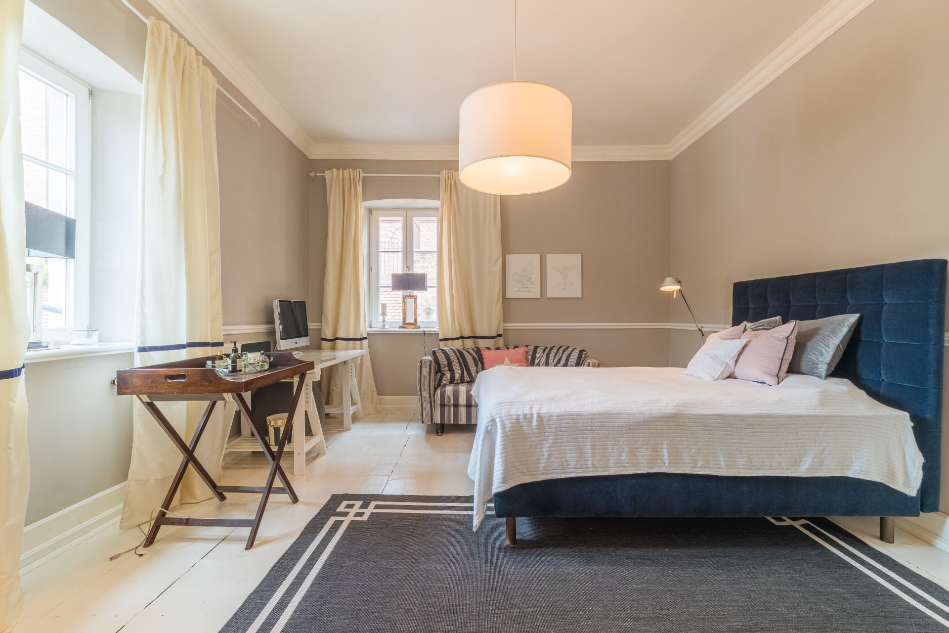Helles Schlafzimmer mit Doppelbett, Schreibtisch, Sessel und großen Fenstern mit Vorhängen.