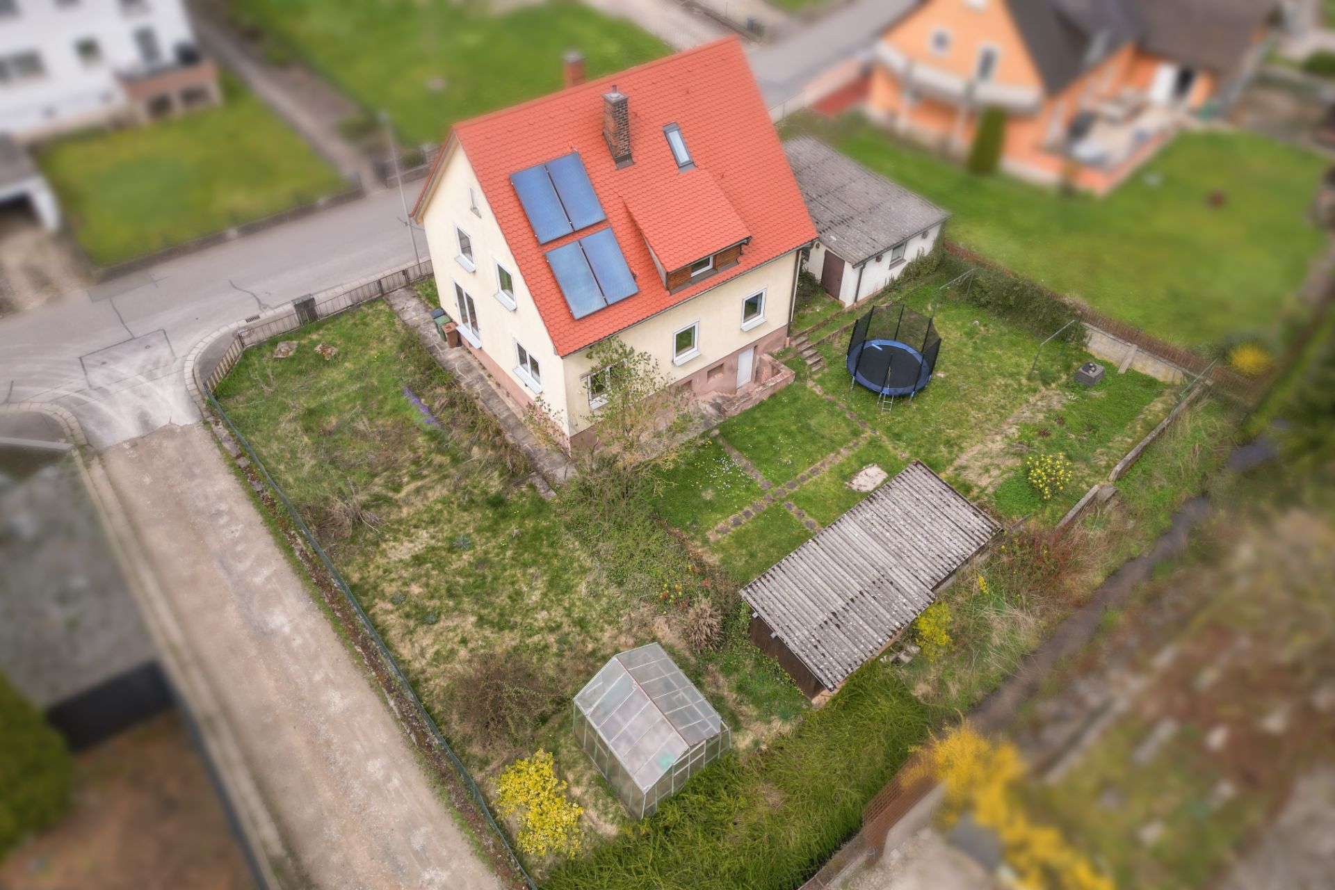 Haus mit rotem Dach, Solarpanels, Garten, Gewächshaus, Schuppen und Trampolin, aus der Vogelperspektive.