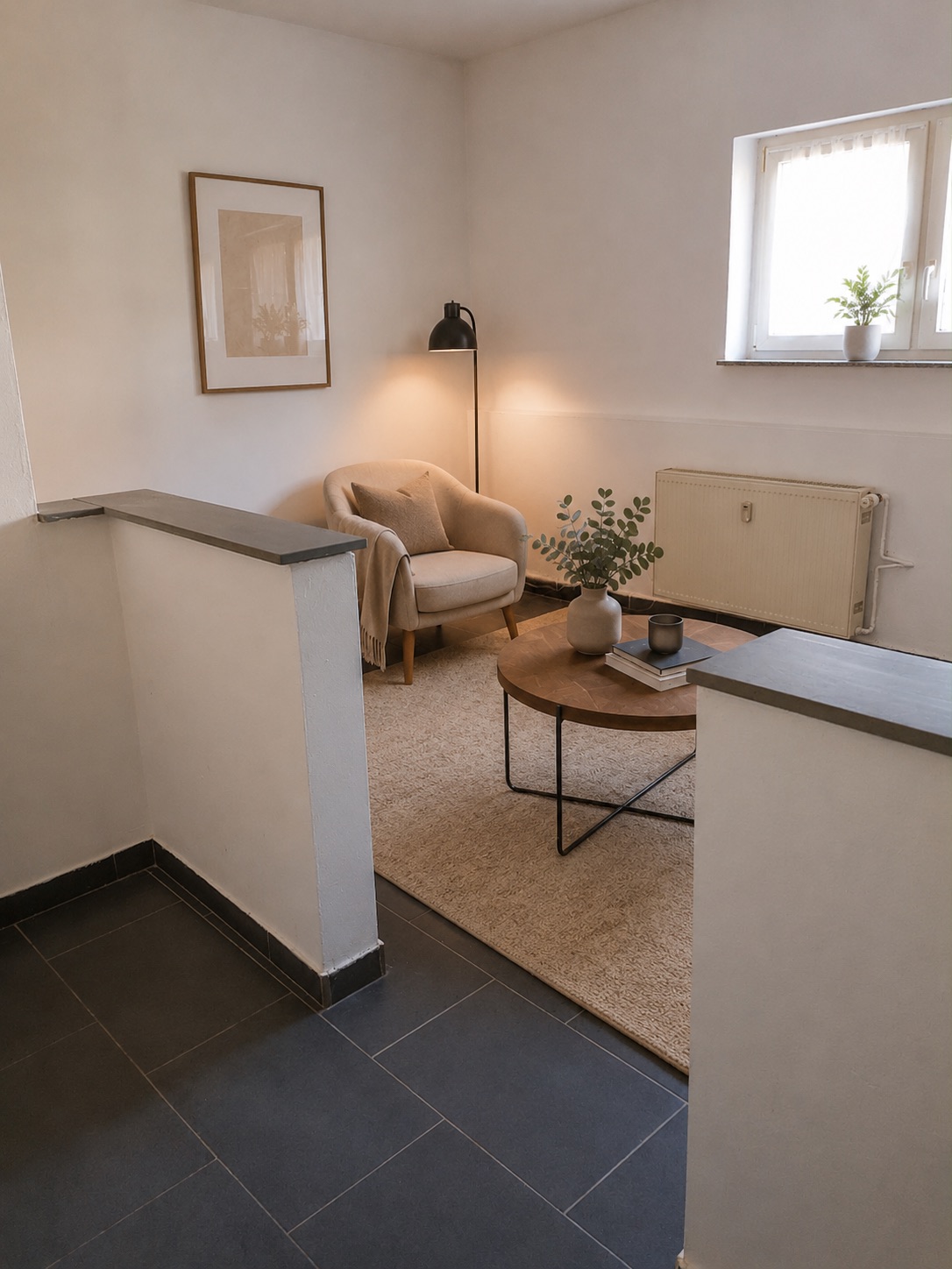 Heller Wohnbereich mit Sessel, Stehlampe, Couchtisch und kleinem Fenster mit Pflanze.