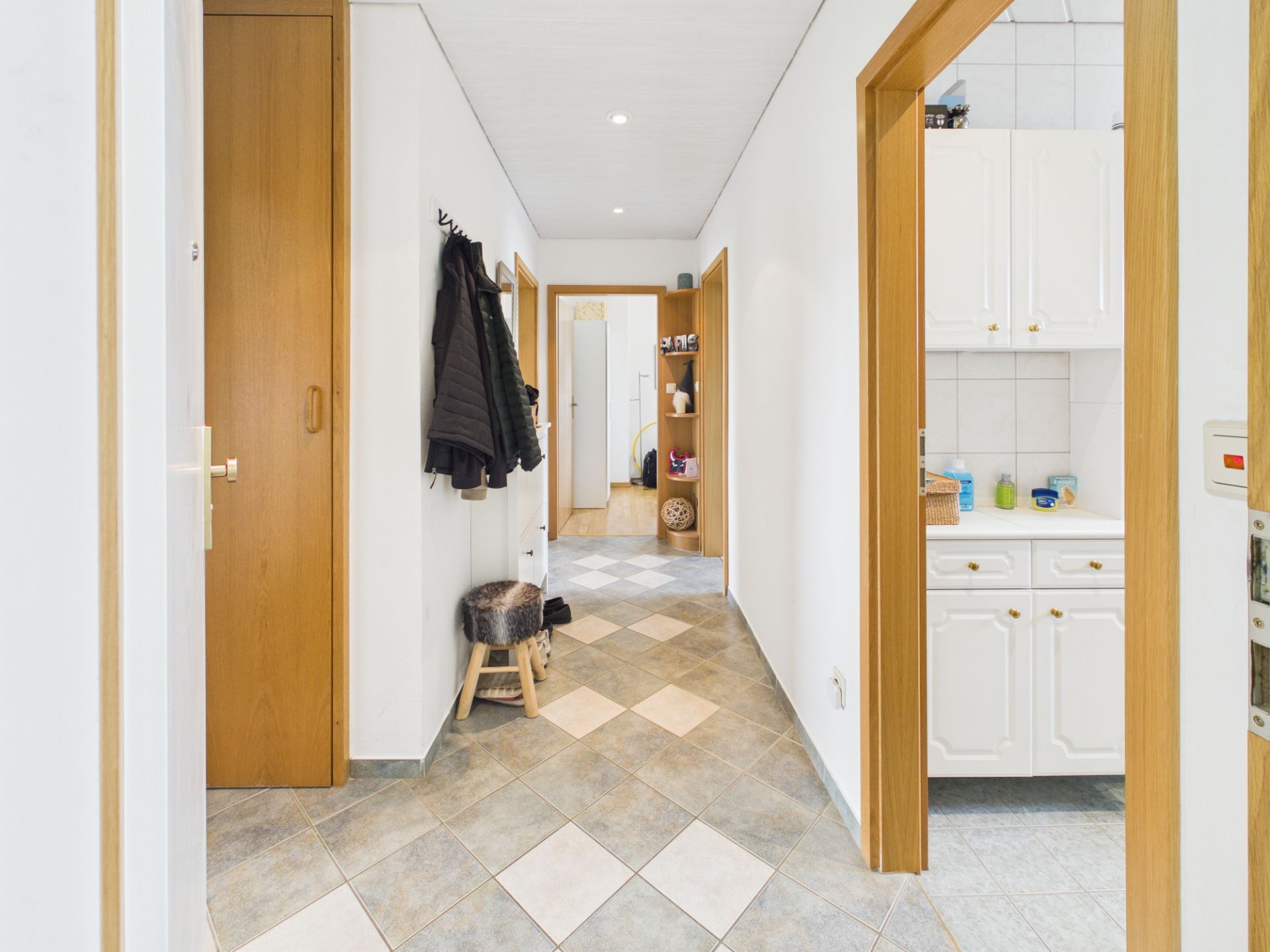 Heller Flur mit Garderobe, Hocker, Fliesenboden und Blick in die Küche und einen weiteren Raum.