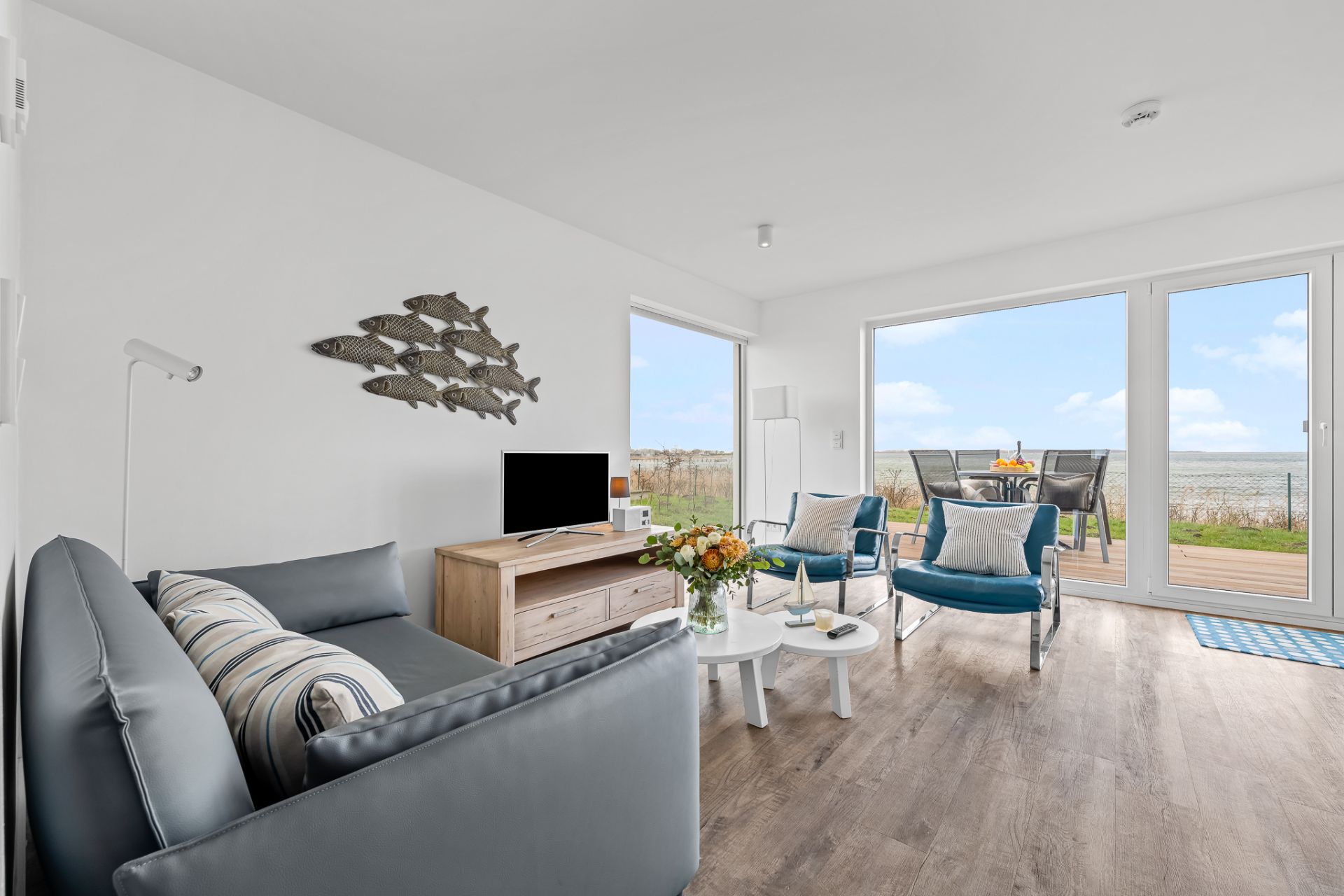 Helles Wohnzimmer mit Sofa, Sessel, Tisch, Fernseher und Meerblick durch große Fenster.