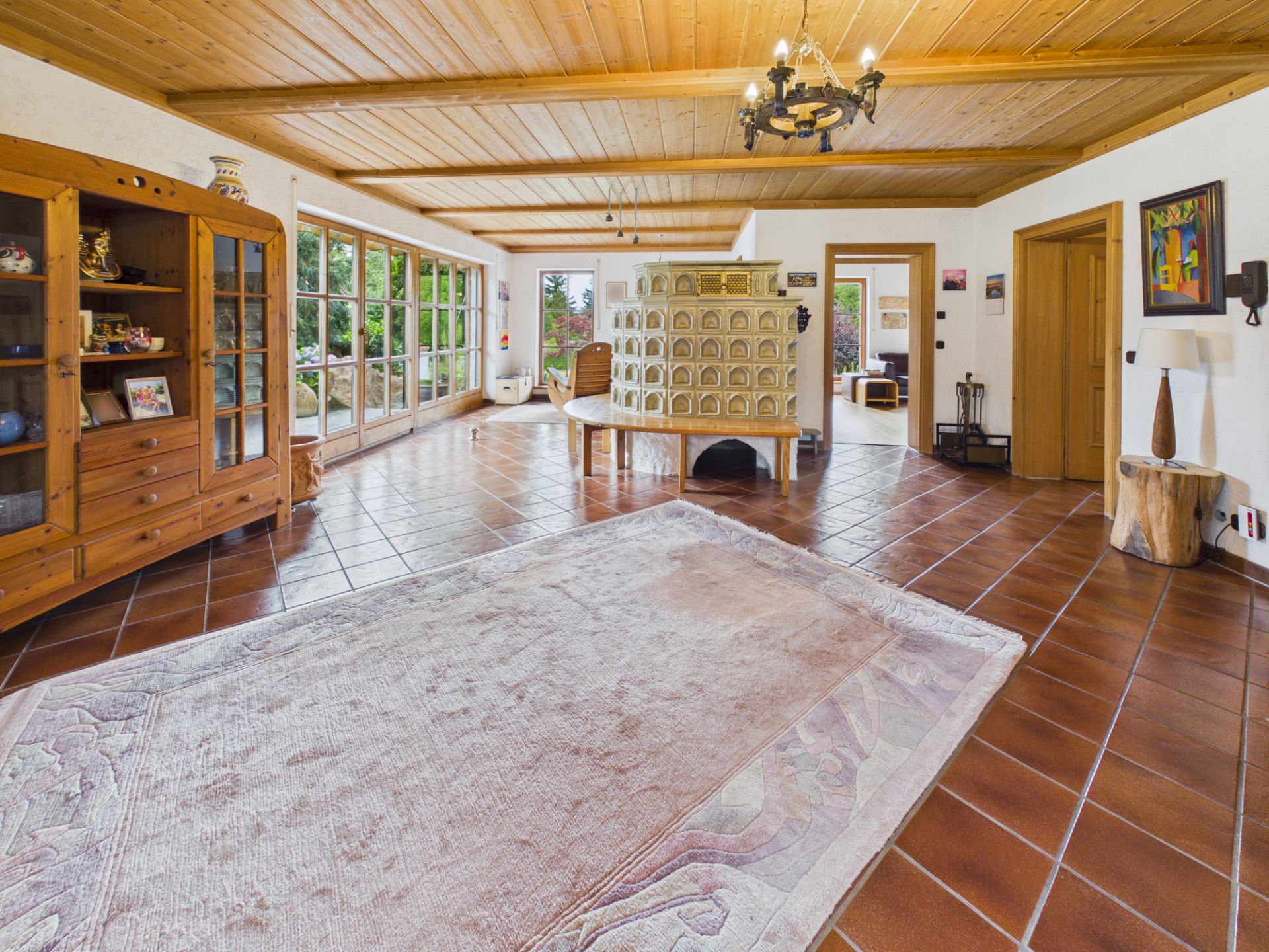 Großes Wohnzimmer mit Fliesenboden, Holzdecke, Kamin und großen Fenstern zum Garten.