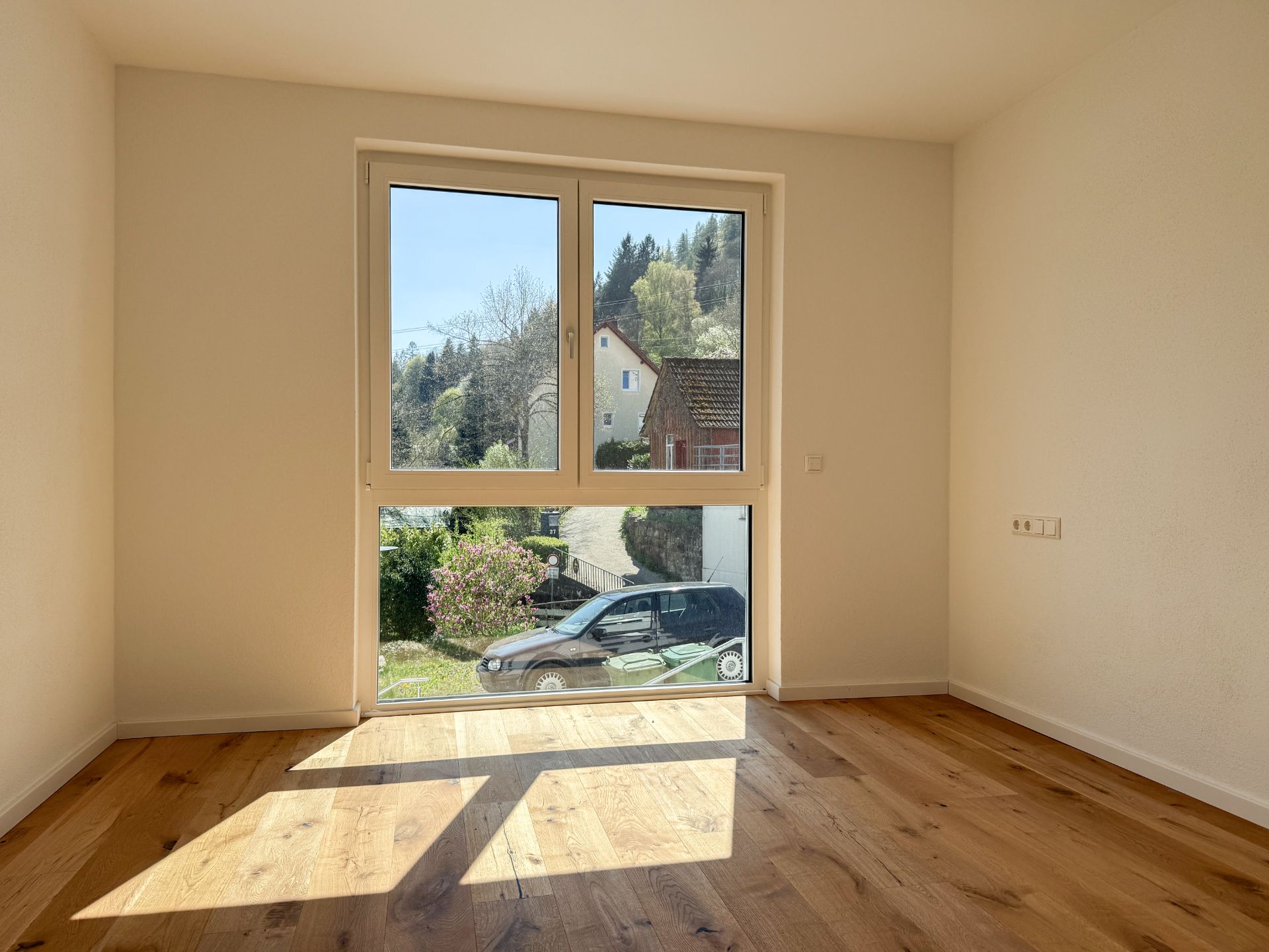 Helles leeres Zimmer mit großem Fenster und Holzfußboden, Blick auf Garten und Auto.
