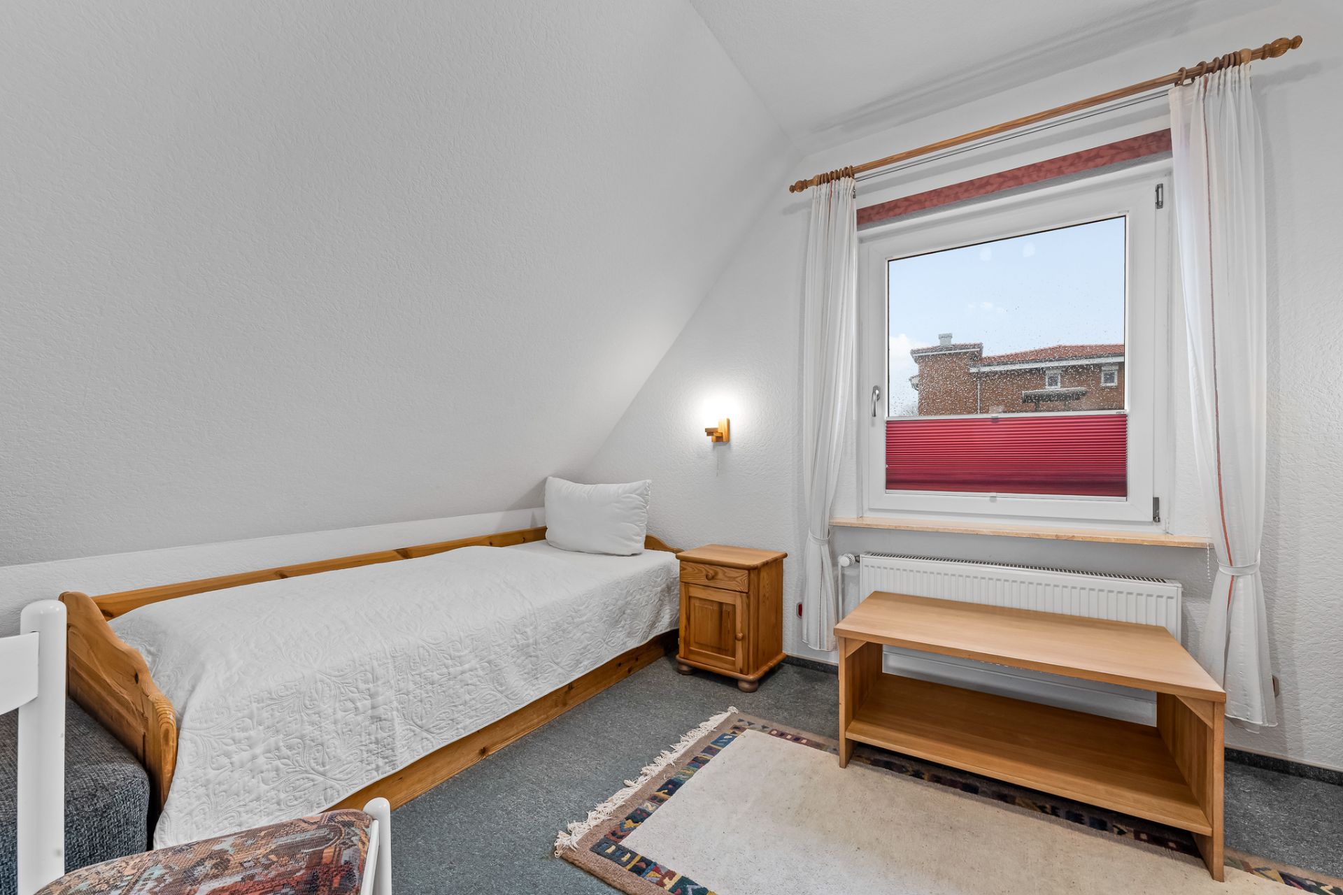 Kleines Schlafzimmer mit Einzelbett, Nachttisch, Holzbank und Fenster mit Vorhängen.