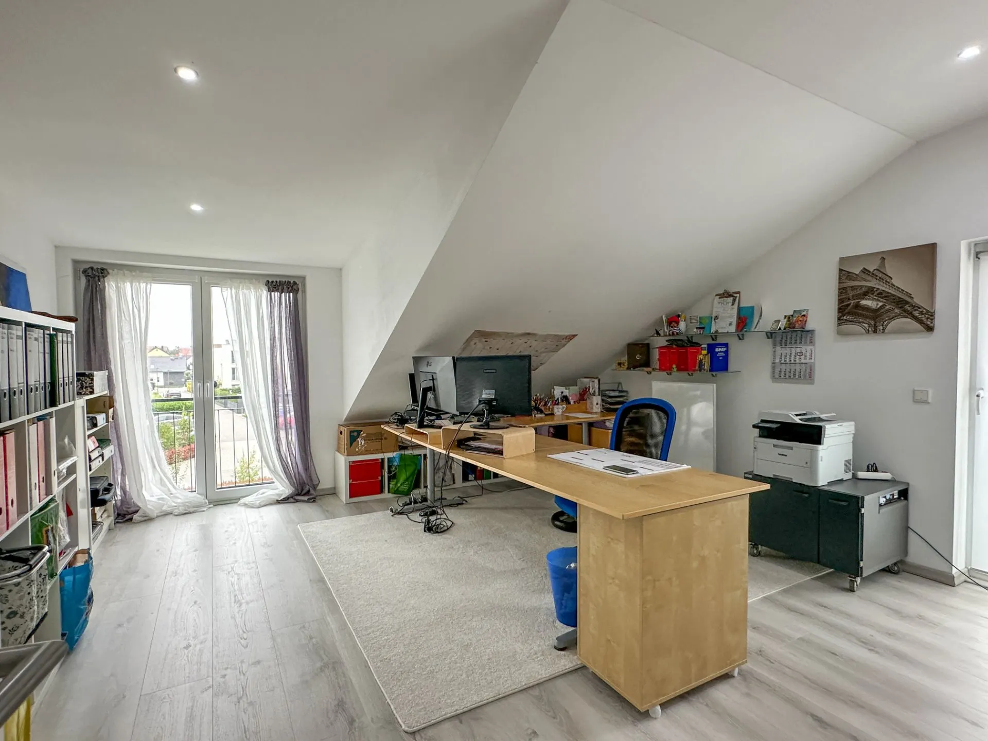 Helles Arbeitszimmer mit Schreibtisch, Computer, Regalen und Balkon­tür unter einer Dachschräge.