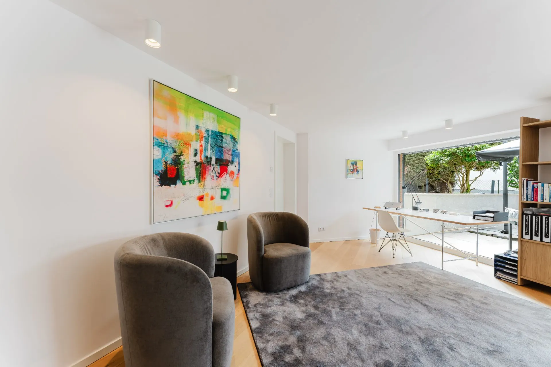 Helles Wohnzimmer mit zwei grauen Sesseln, Teppich, Schreibtisch und großem Fenster zum Garten.