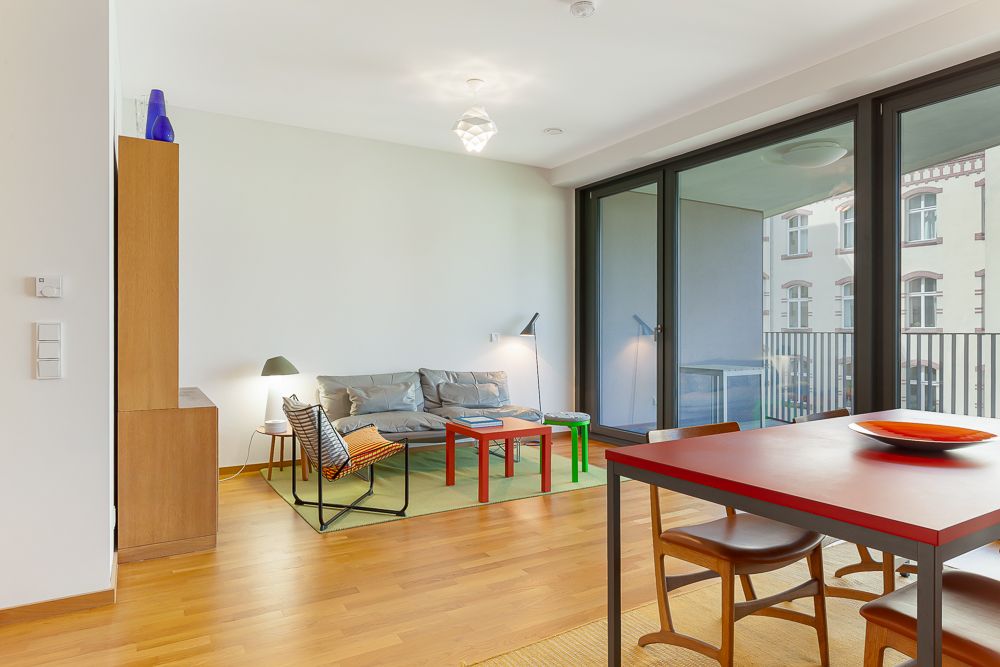 Modernes Wohnzimmer mit Sofa, bunten Tischen, Esstisch und Zugang zu einem Balkon durch Glas-Schiebetüren.