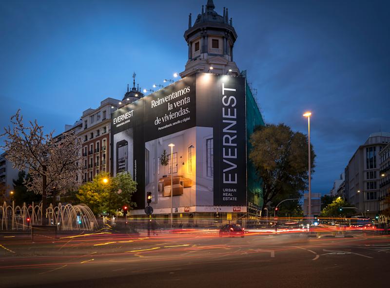 Edificio residencial en una esquina de calle de noche con gran cartel inmobiliario en la fachada.