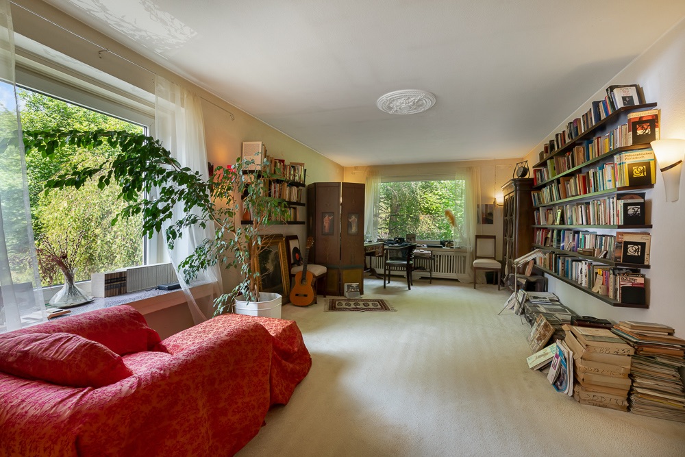 Helles Wohnzimmer mit rotem Sofa, großen Fenstern, Bücherregalen und Zimmerpflanzen.
