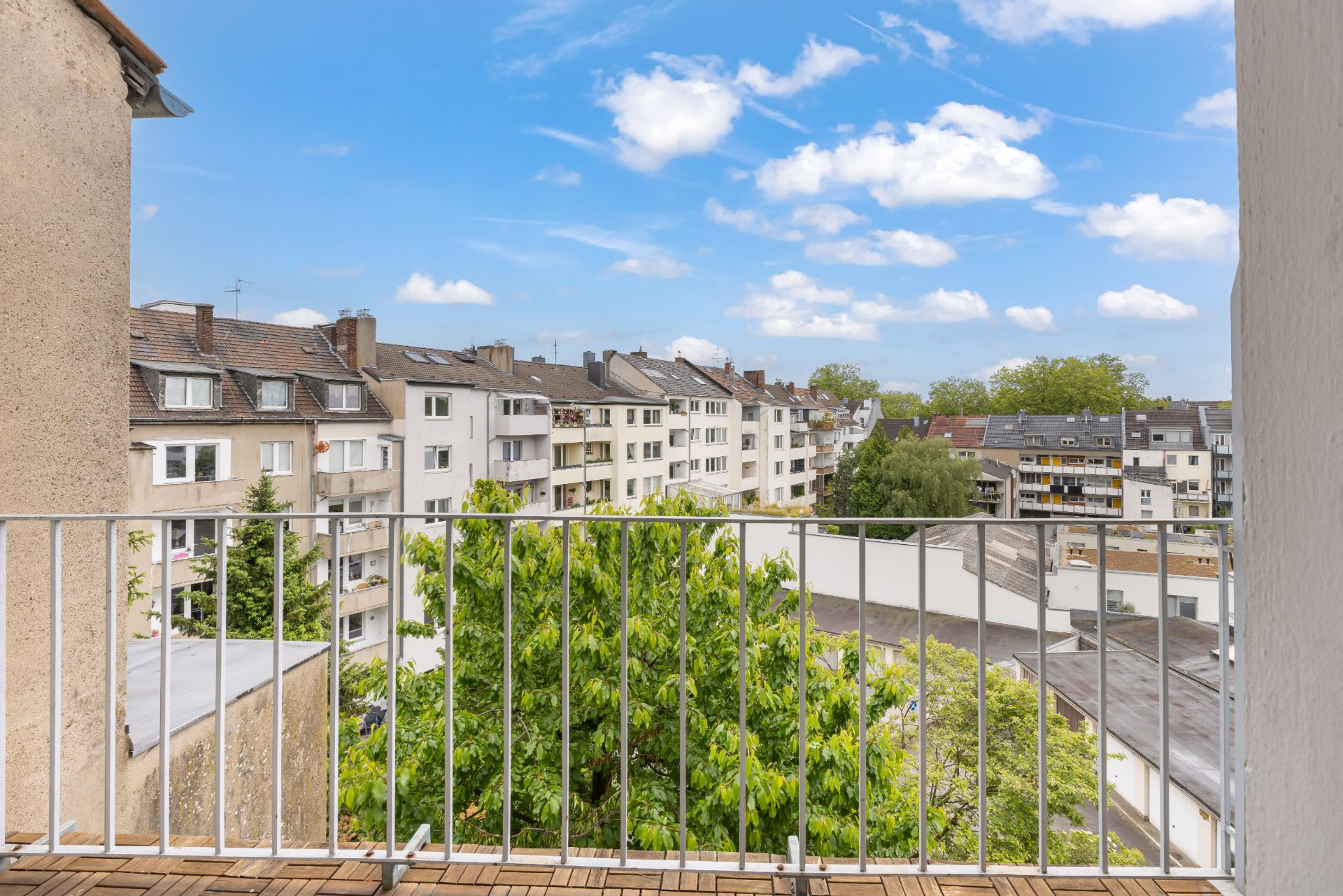 Blick von einem Balkon auf Wohnhäuser, Bäume und blauen Himmel mit Wolken.