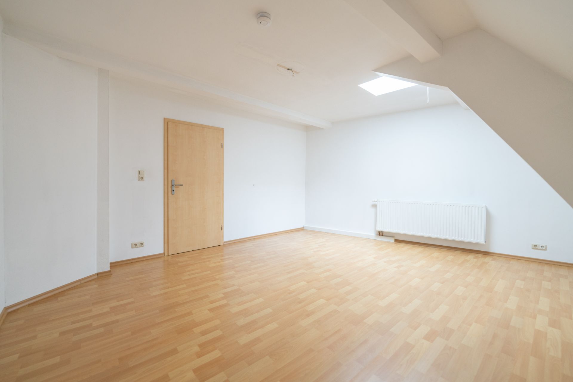 Leerer Raum mit hellem Holzboden, weißer Wand, Tür und Heizkörper unter einem Fenster.
