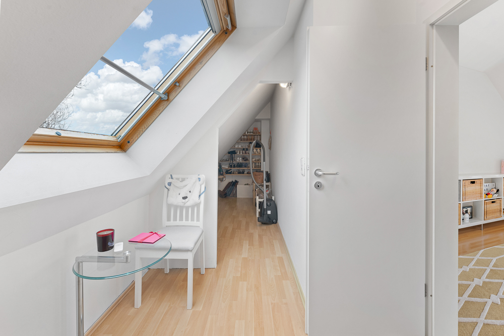 Helle Dachgeschossflur mit großem Fenster, Stuhl, Glastisch und Holzboden.