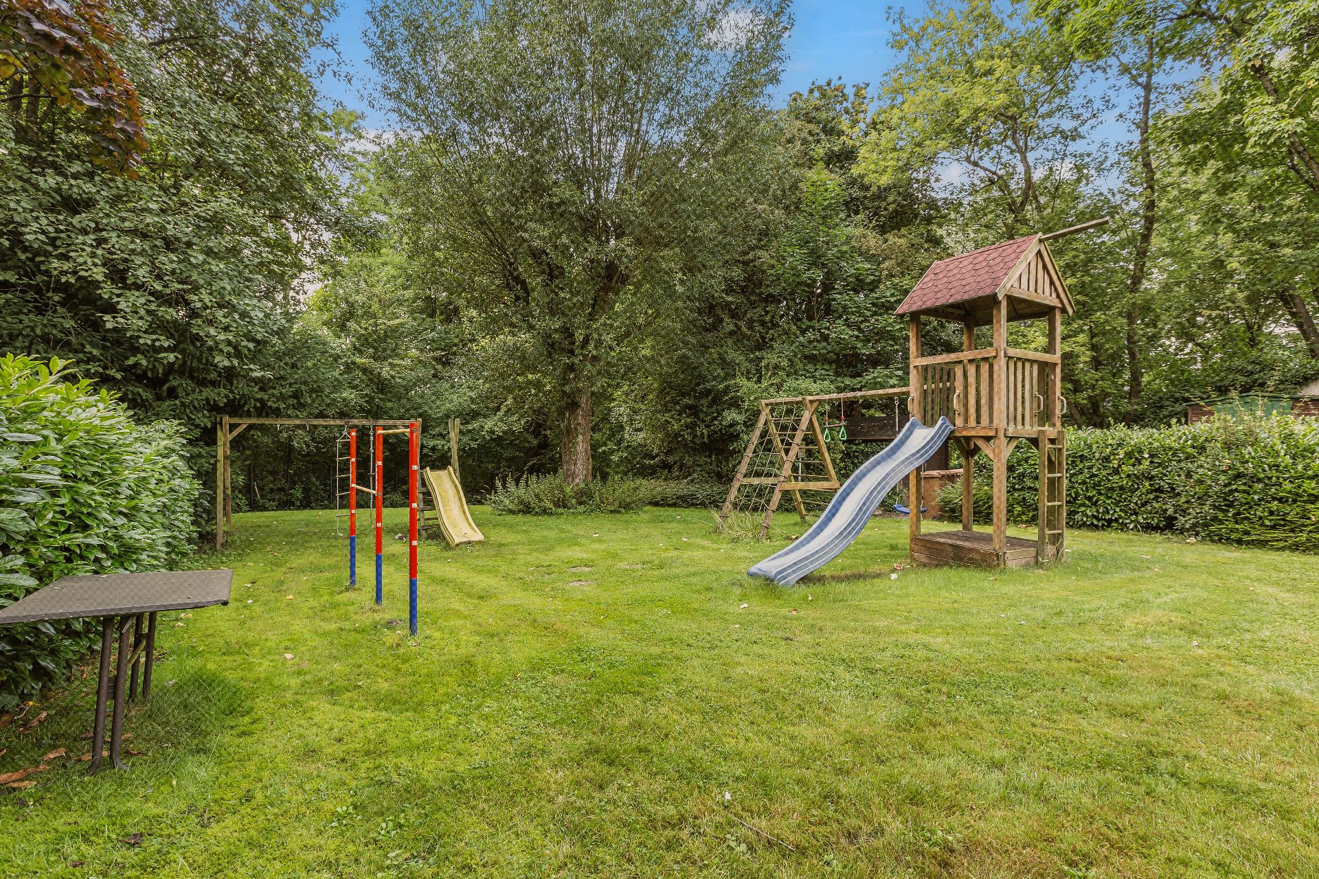 Garten mit Spielplatz, Rutsche, Klettergerüst und Schaukel, umgeben von Bäumen und Rasen.
