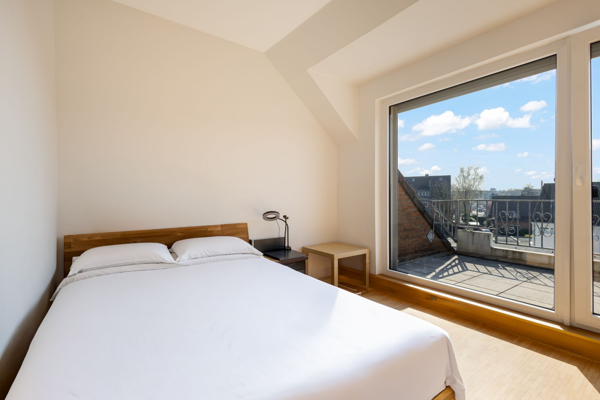 Helles Schlafzimmer mit Doppelbett, Nachttisch und großem Fenster mit Blick auf einen Balkon.