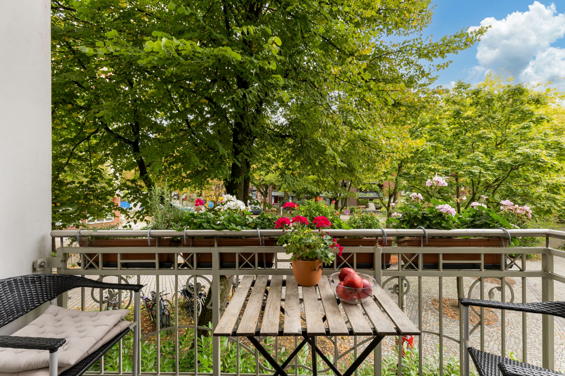 Kleiner Balkon mit Holztisch, zwei Stühlen und Blumen, umgeben von grünen Bäumen.