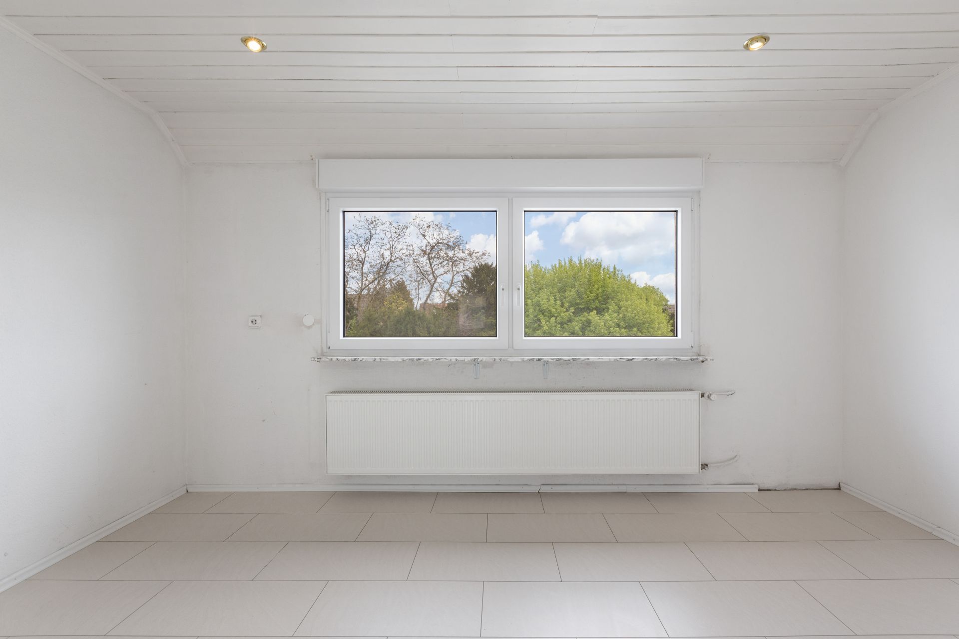 Heller, leerer Raum mit großem Fenster, weißer Heizung und Fliesenboden.