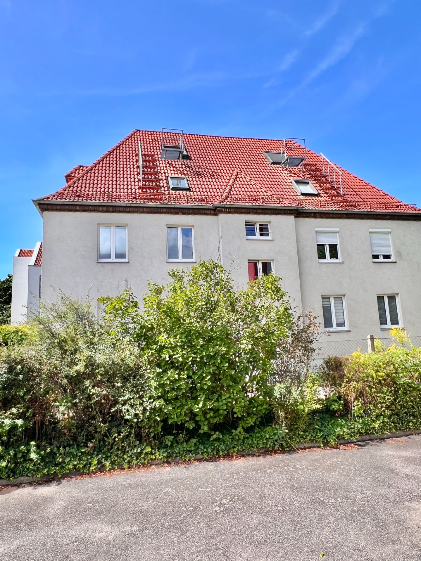 Mehrfamilienhaus mit rotem Dach, mehreren Fenstern und Vorgarten mit Büschen.