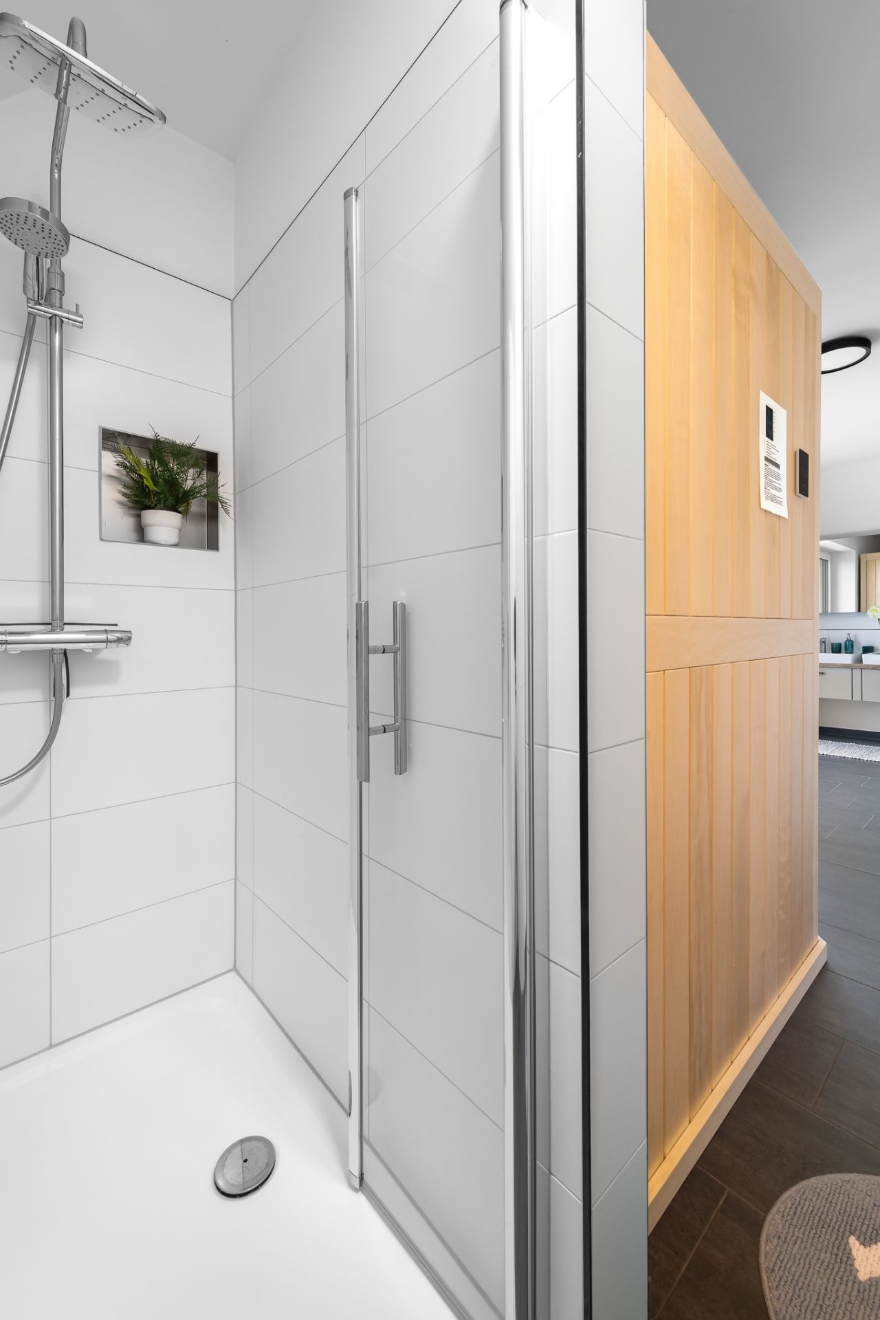 Weiße Dusche mit Glaswand und eingebautem Regal mit Pflanze, daneben eine Holzsauna.