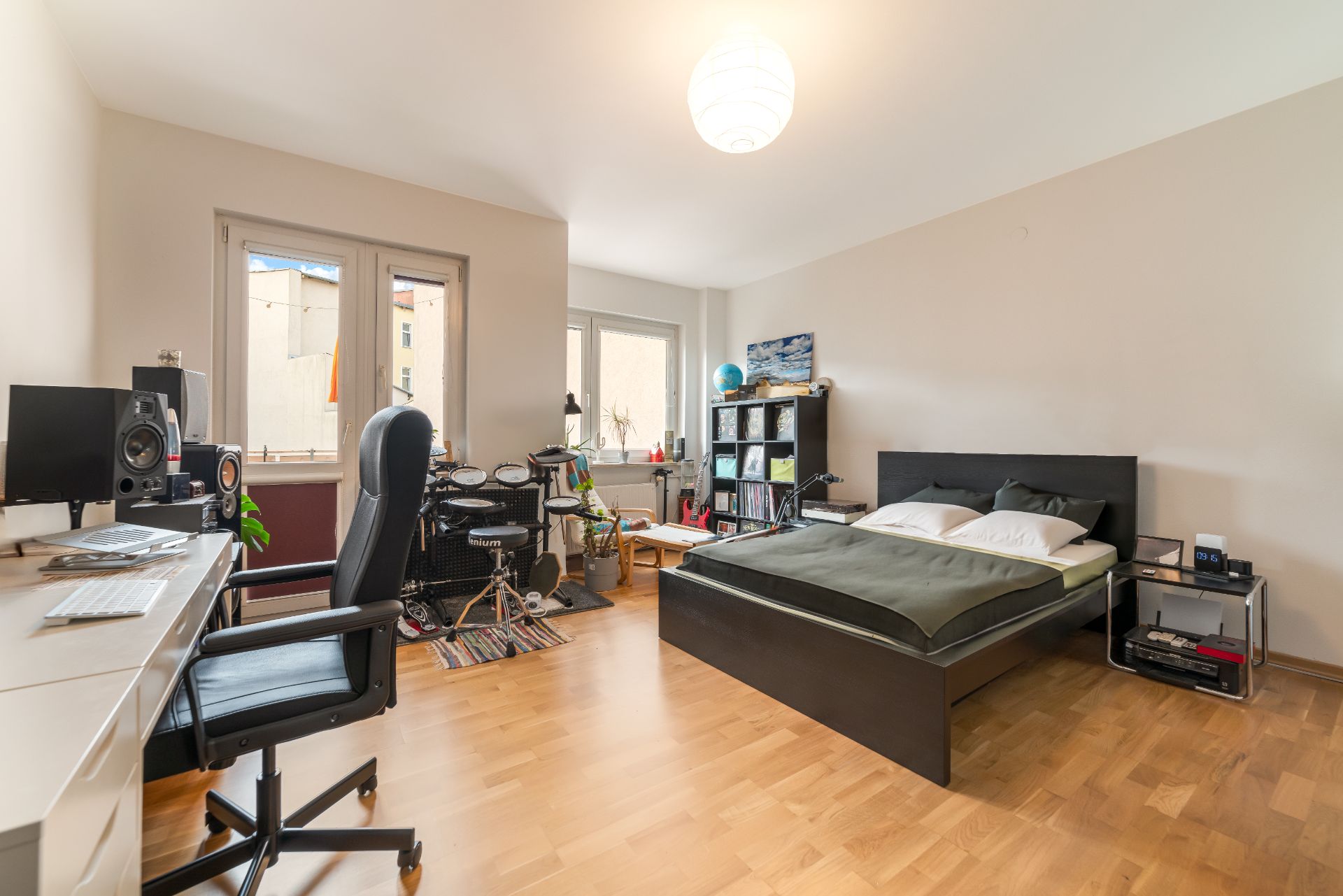 Modernes Schlafzimmer mit Schreibtisch, Doppelbett, Regalen und E-Drum-Set auf Holzfußboden.