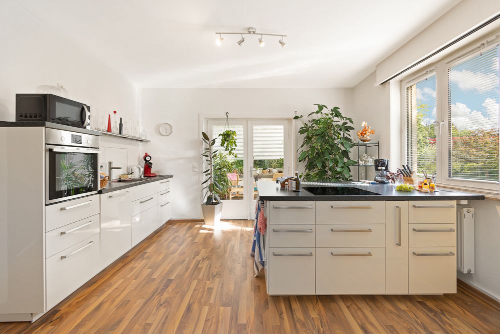 Helle, moderne Küche mit weißen Schränken, Holzboden, großen Fenstern und Pflanzen.