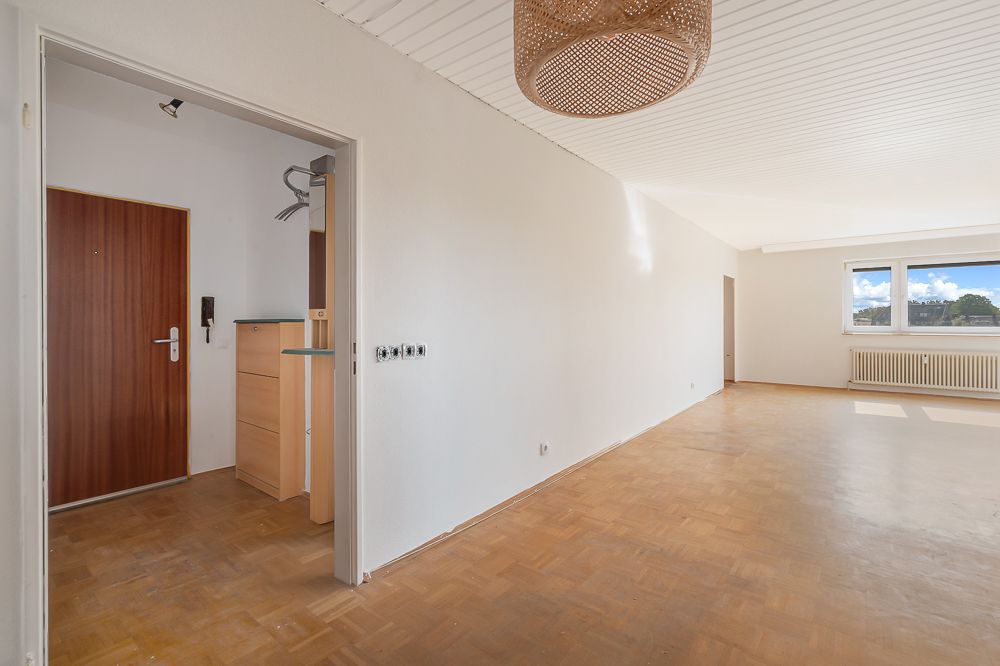 Heller leerer Wohnraum mit Holzboden, großem Fenster und Blick in den Eingangsbereich.