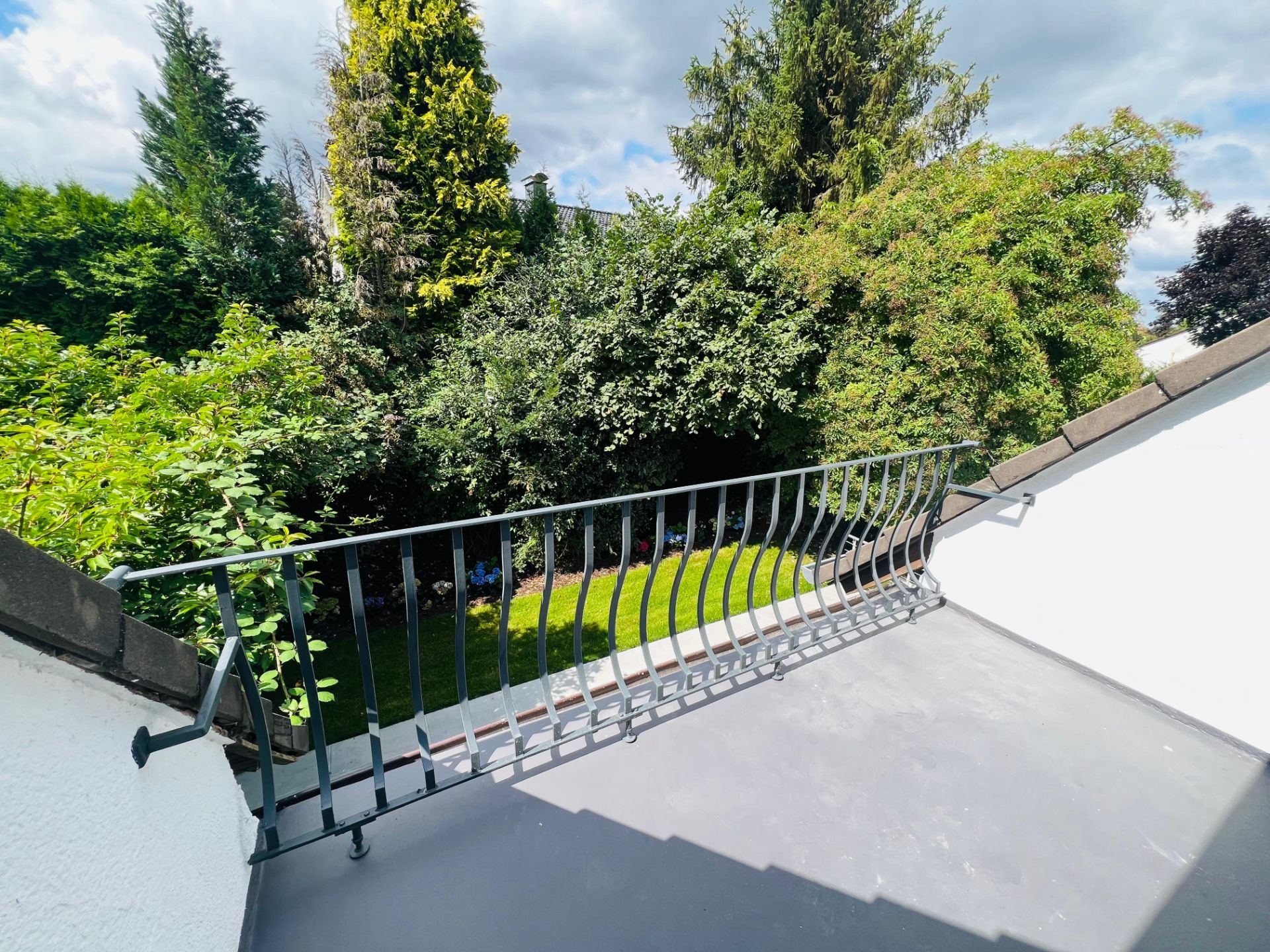 Kleiner Balkon mit Metallgeländer und Blick auf einen grünen Garten mit Bäumen.