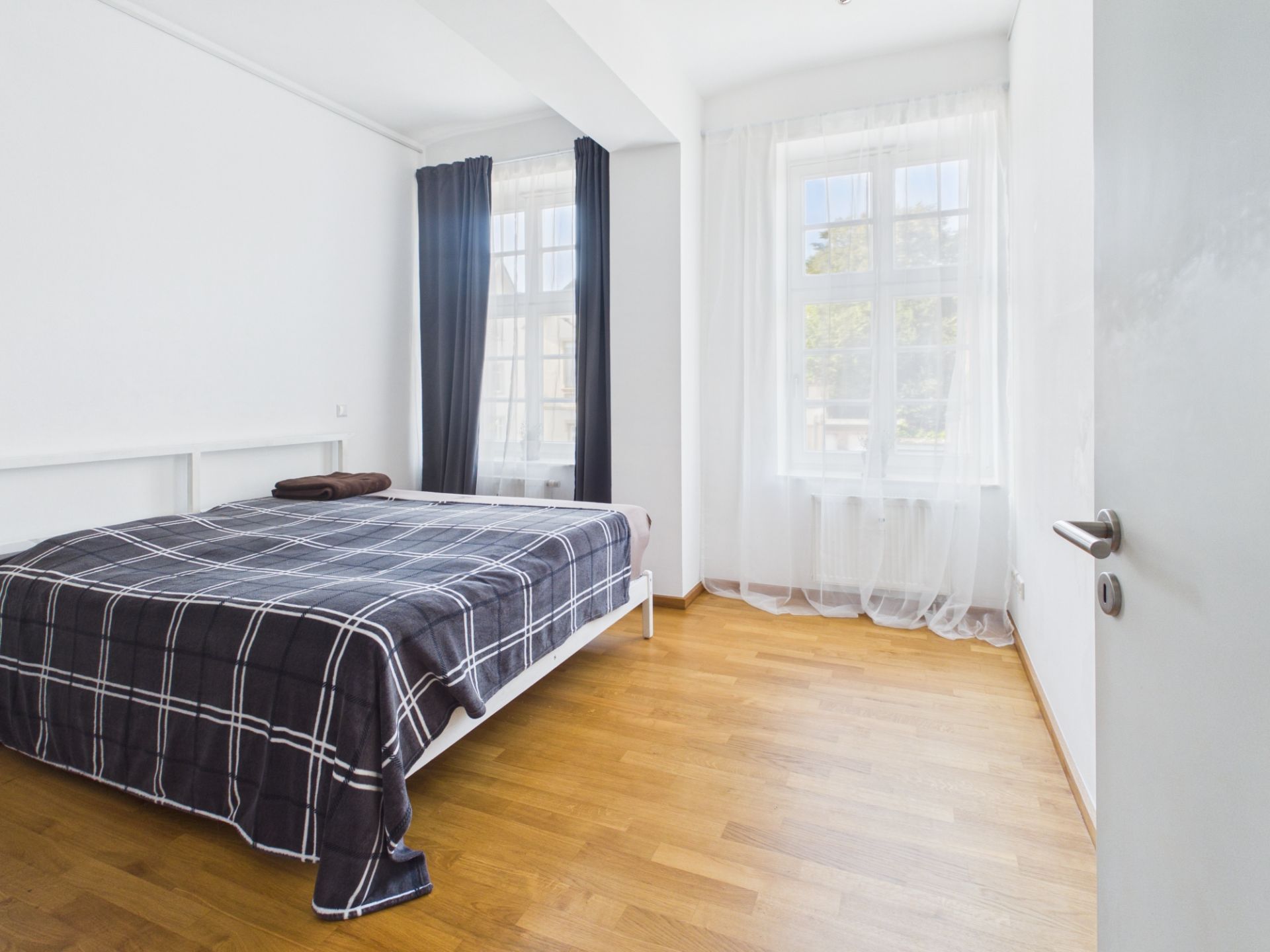 Helles Schlafzimmer mit Holzboden, Bett und zwei Fenstern mit Vorhängen.