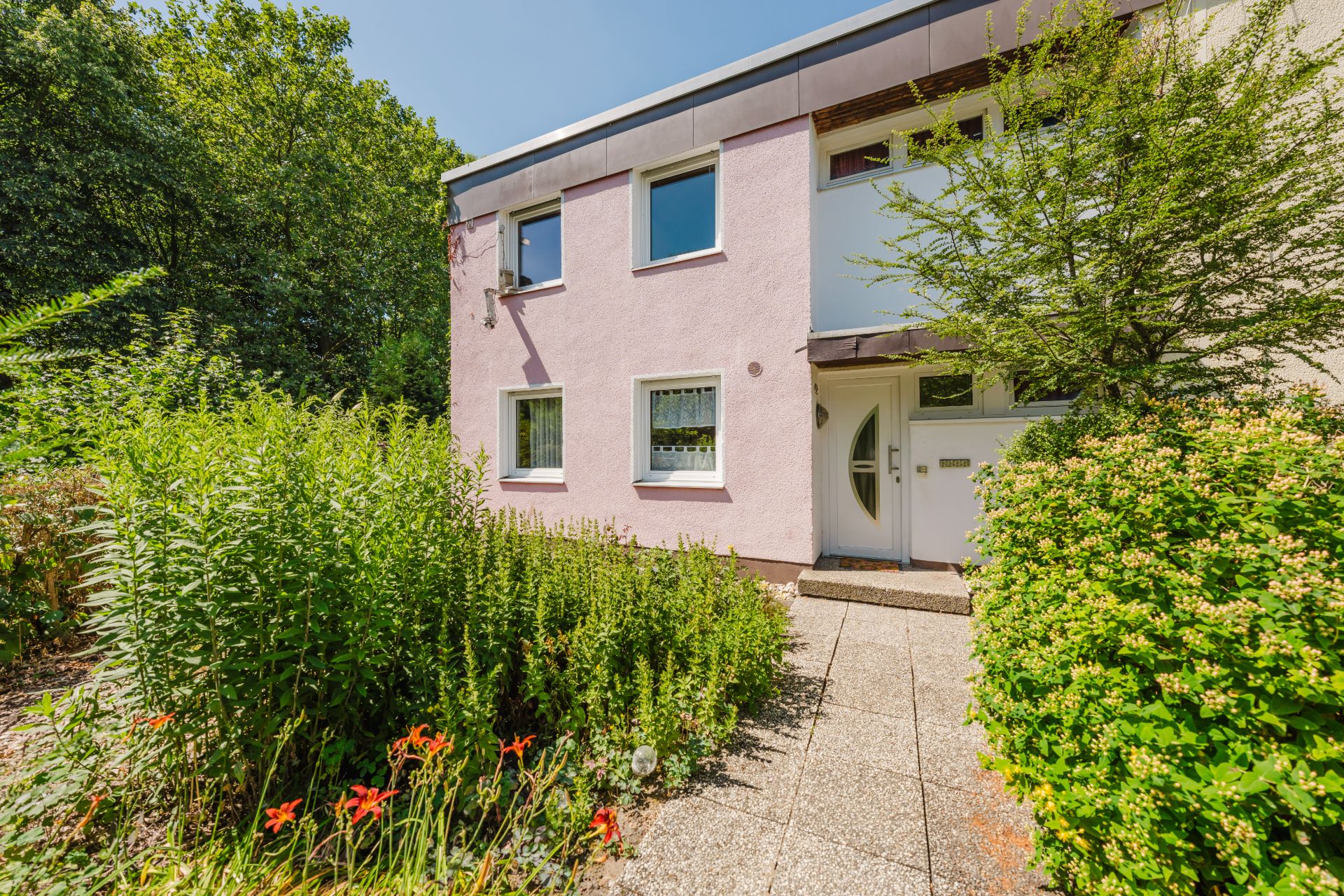 Zweistöckiges Haus mit rosa Fassade, großen Fenstern und Gartenweg, umgeben von viel Grün.