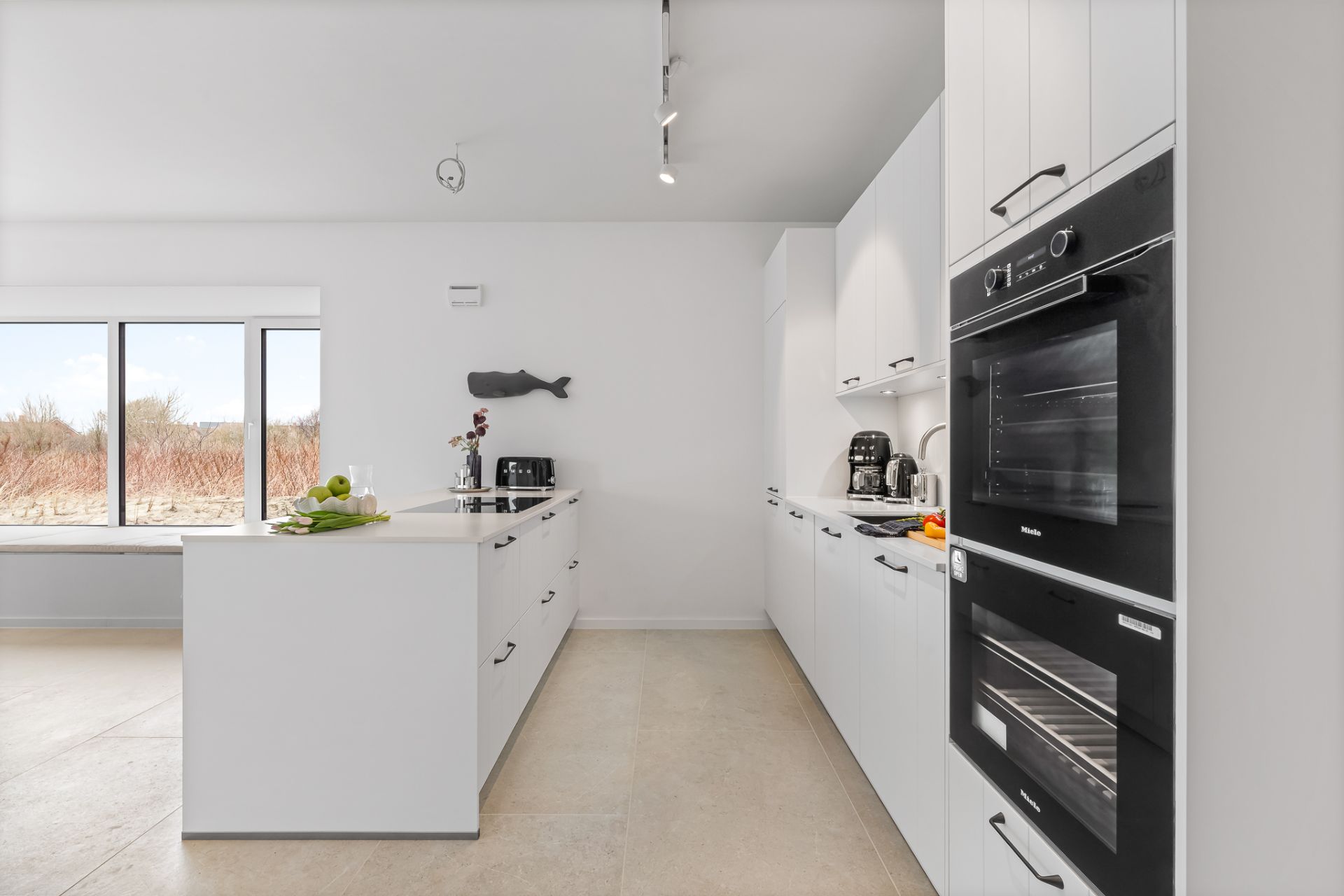 Moderne, weiße Einbauküche mit Kochinsel, Backofen, Fenstern und Küchengeräten.