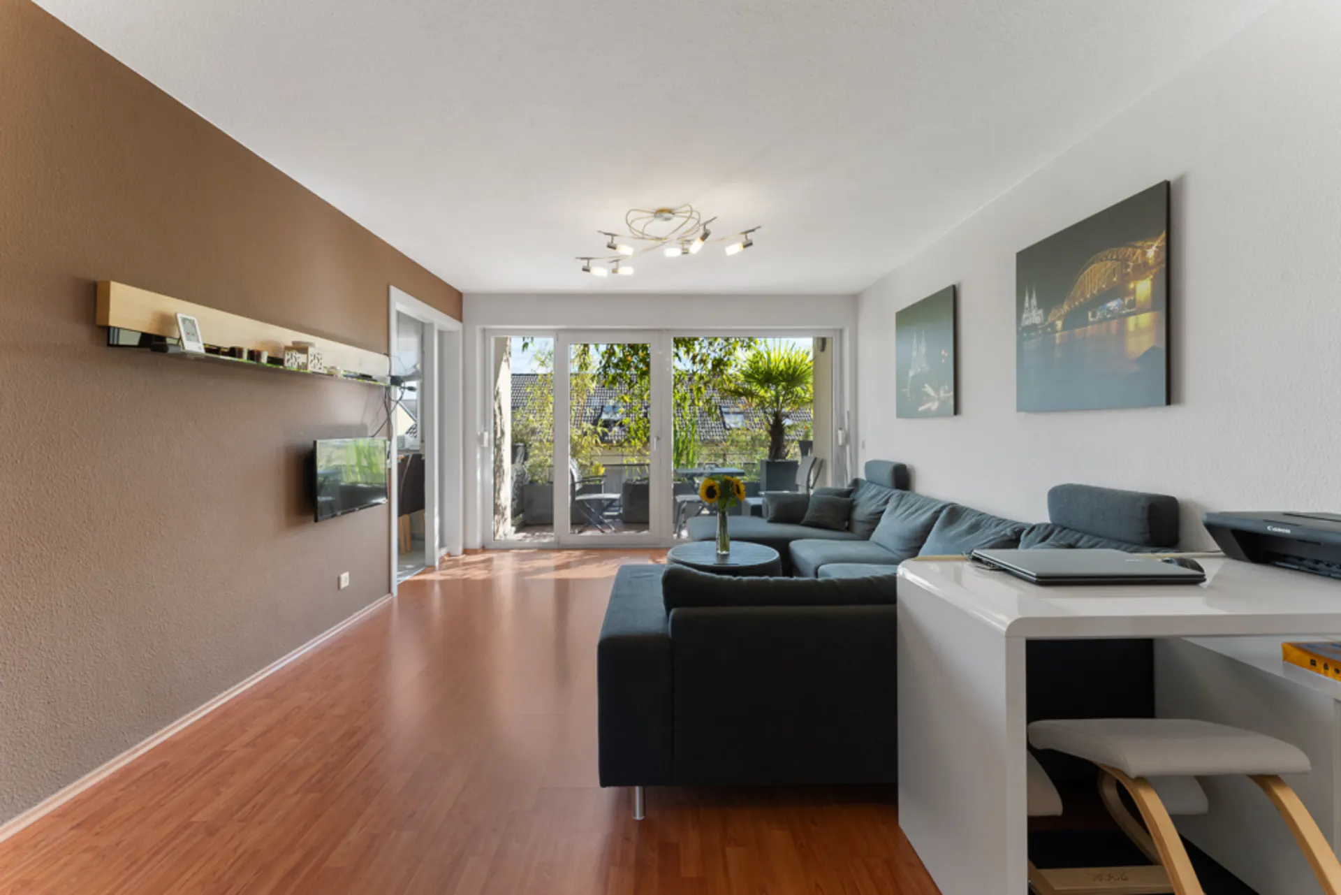 Modernes Wohnzimmer mit Ecksofa, Fernseher, Schreibtisch und Zugang zu einem Balkon mit Pflanzen.