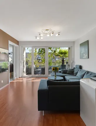 Modernes Wohnzimmer mit Ecksofa, Fernseher, Schreibtisch und Zugang zu einem Balkon mit Pflanzen.