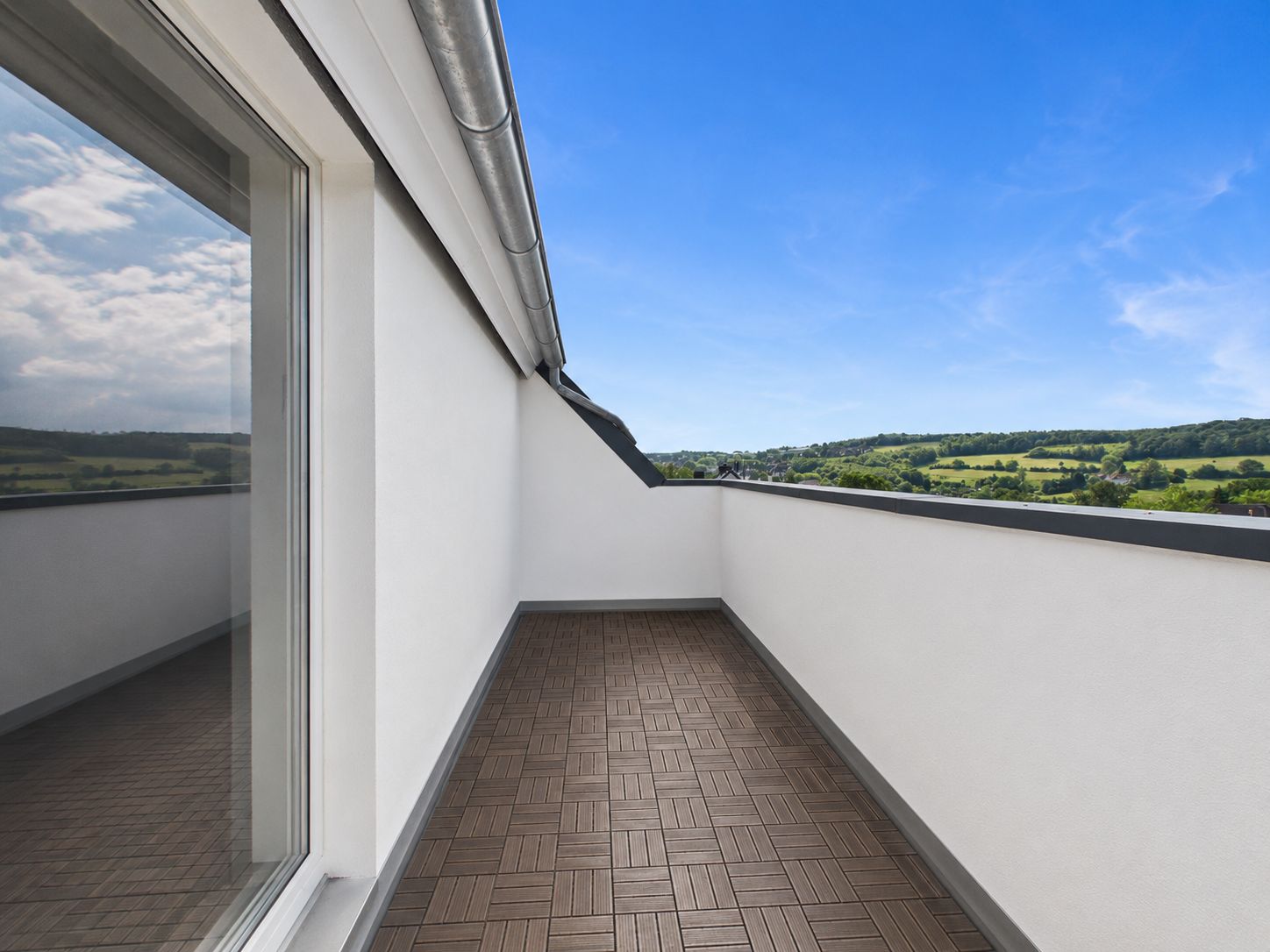 Balkon mit braunem Bodenbelag und Ausblick auf grüne Landschaft unter blauem Himmel.