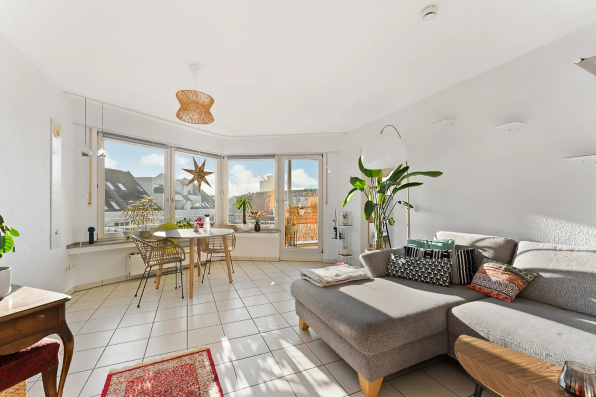 Helles Wohnzimmer mit grauem Sofa, Esstisch am Fenster und mehreren Pflanzen.
