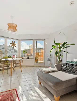 Helles Wohnzimmer mit grauem Sofa, Esstisch am Fenster und mehreren Pflanzen.