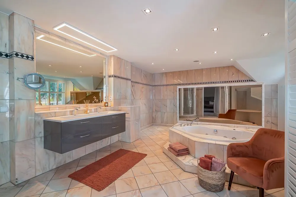 Großes Badezimmer mit Eckbadewanne, Doppelwaschbecken, Spiegel und einem Sessel.
