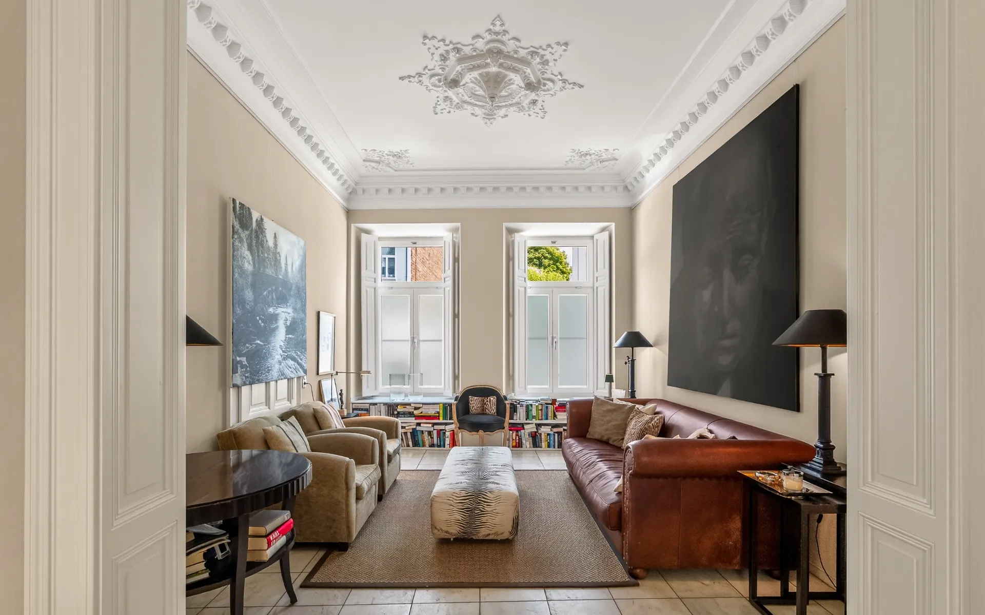 Helles Wohnzimmer mit Sofas, Sesseln, großem Teppich, Kunst an den Wänden und Bücherregalen vor zwei Fenstern.