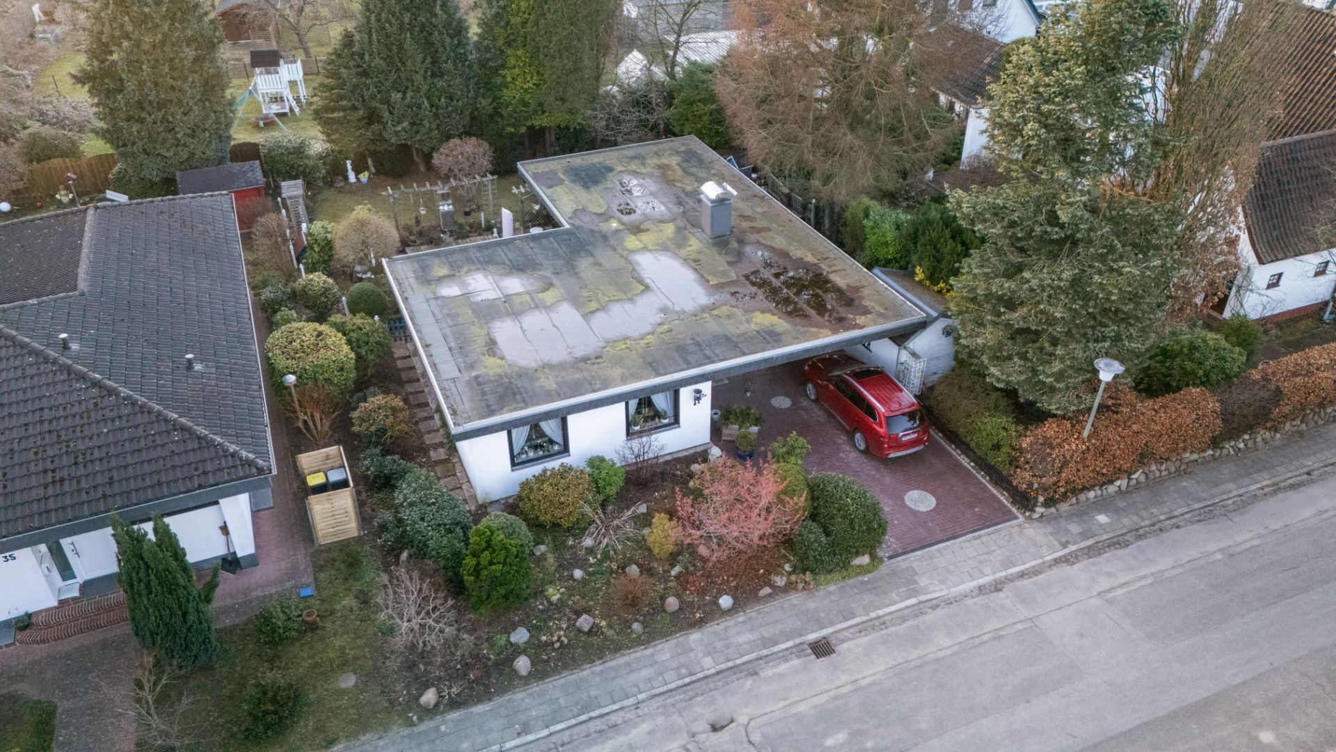 Luftaufnahme eines Bungalows mit Flachdach, Garten und rotem Auto im Carport.
