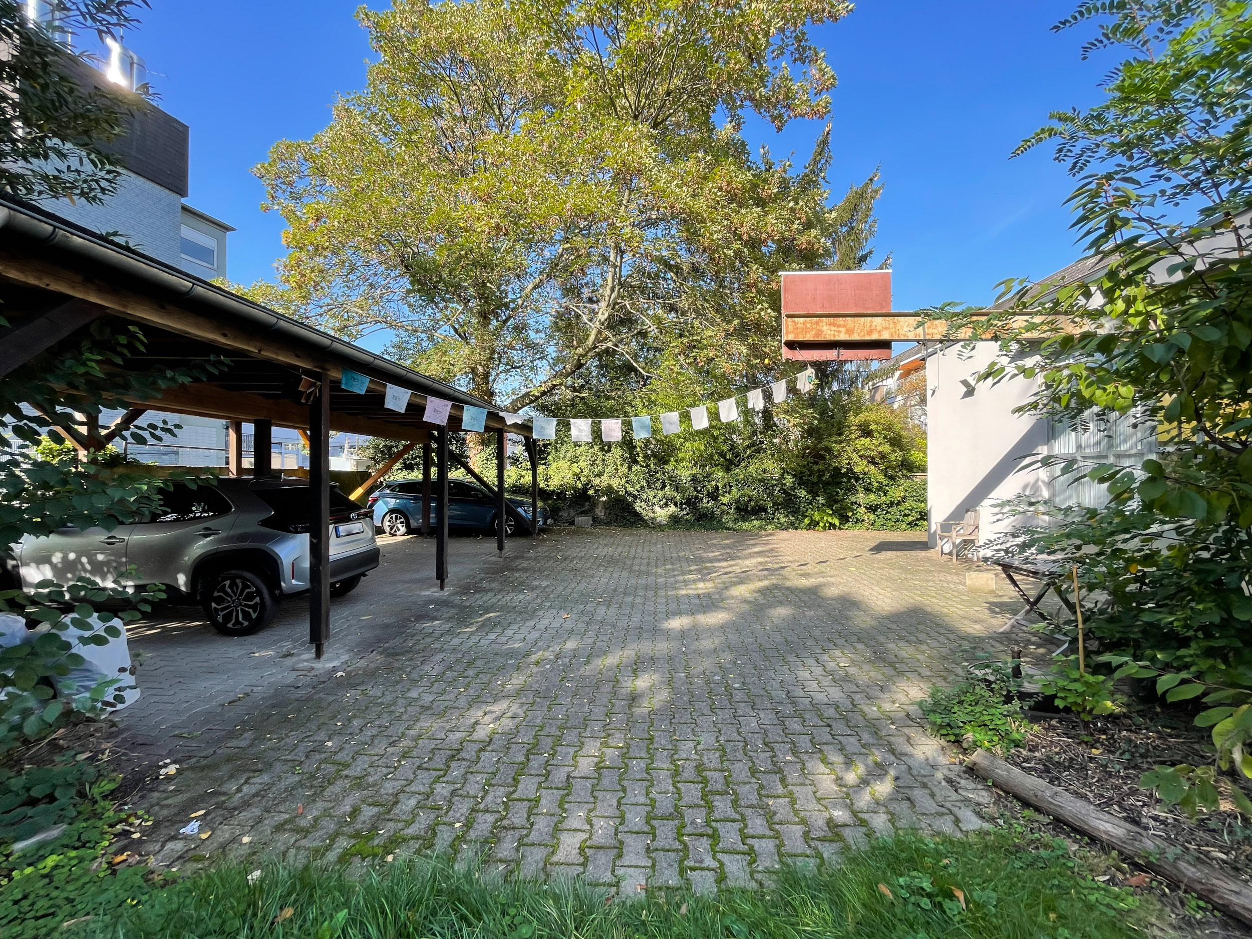 Offener Carport mit zwei geparkten Autos, Pflastersteine, bunte Wimpel und Bäume im Hintergrund.