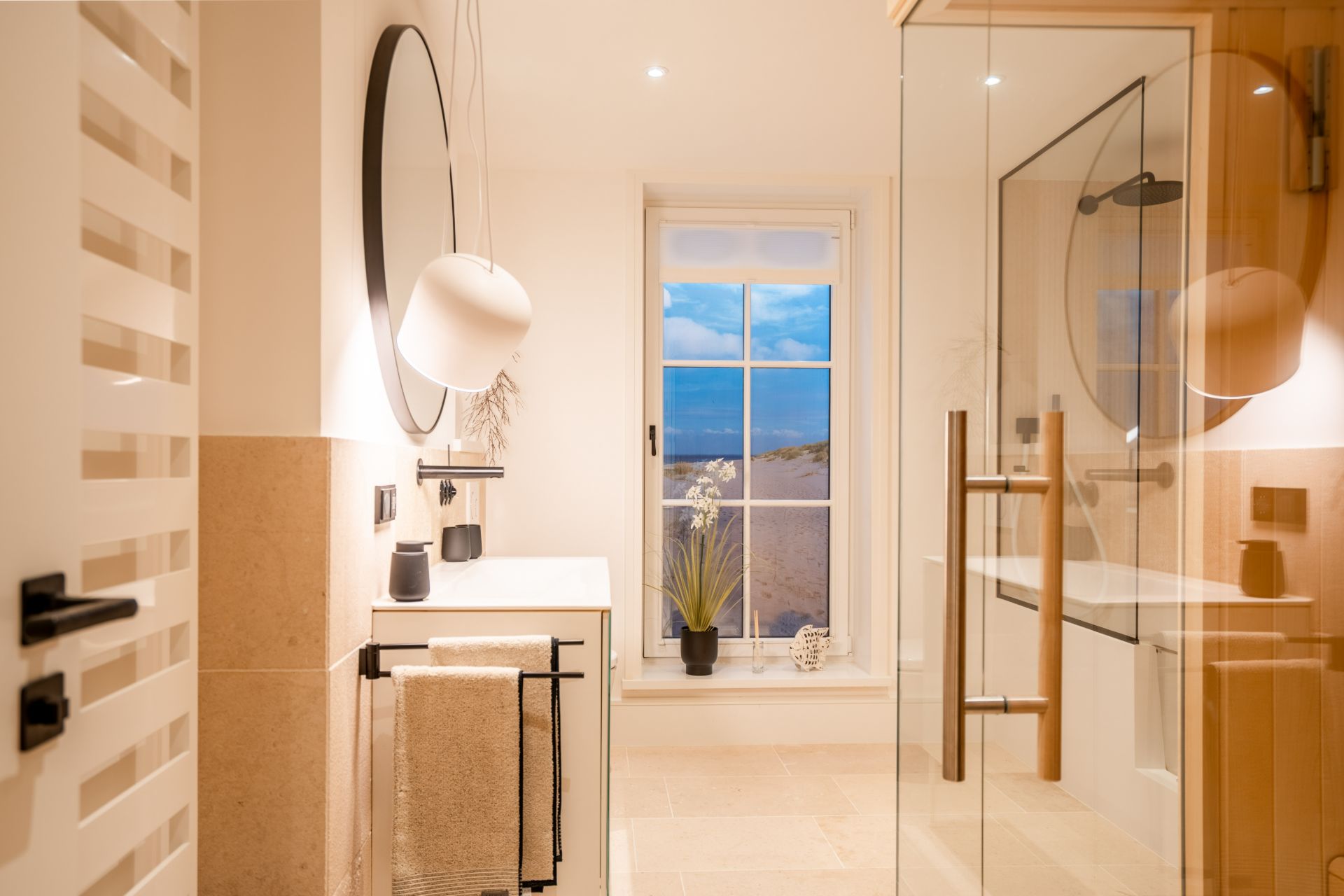 Modernes Badezimmer mit großem Fenster, runder Spiegel, Dusche und hellen Fliesen.