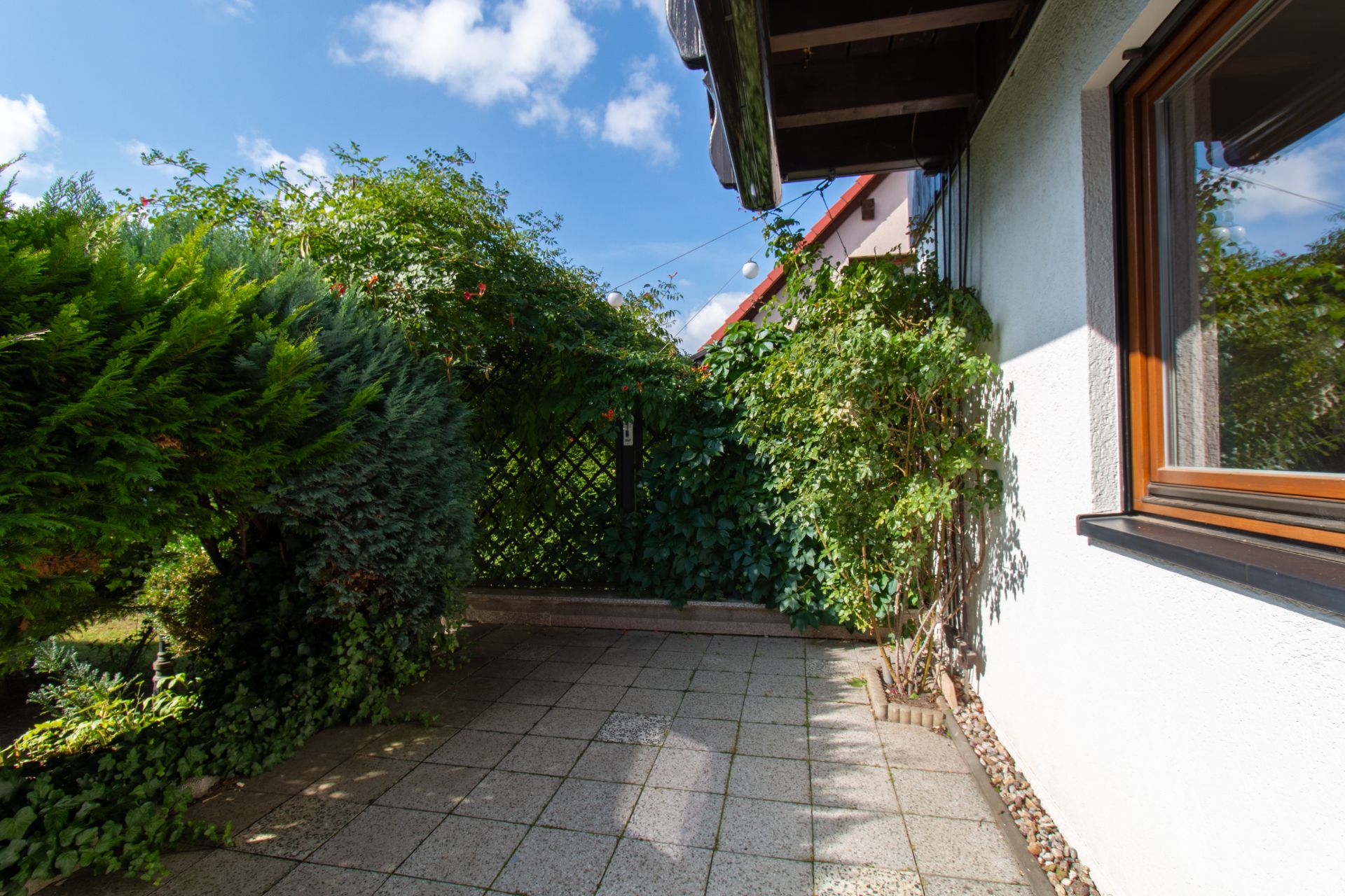 Kleine Terrasse mit Steinplatten neben einem weißen Haus, umgeben von grünen Büschen.