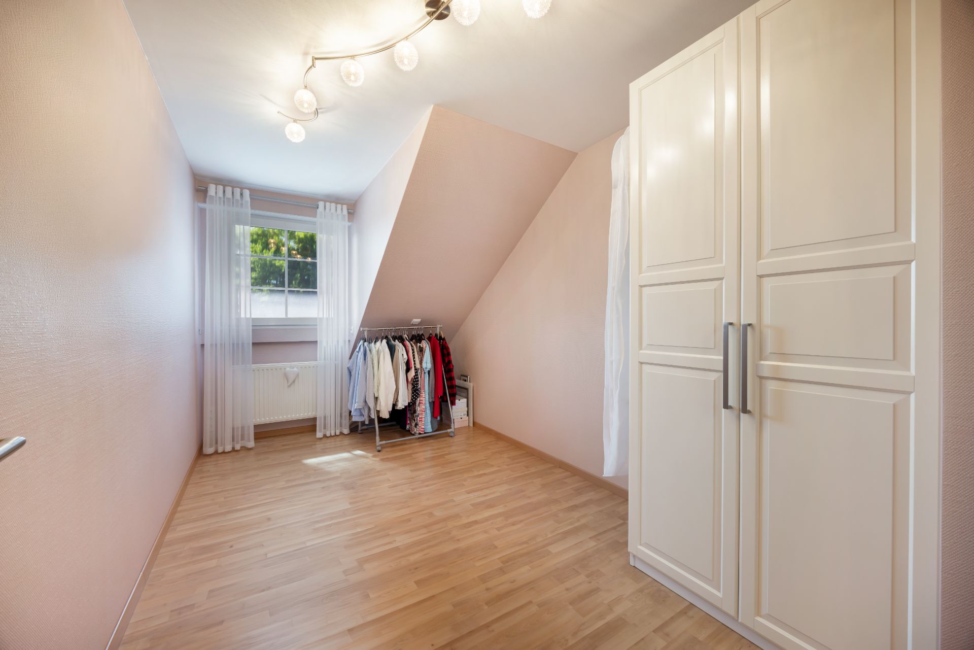 Heller Raum mit Kleiderschrank, Garderobe und Fenster mit weißen Vorhängen.