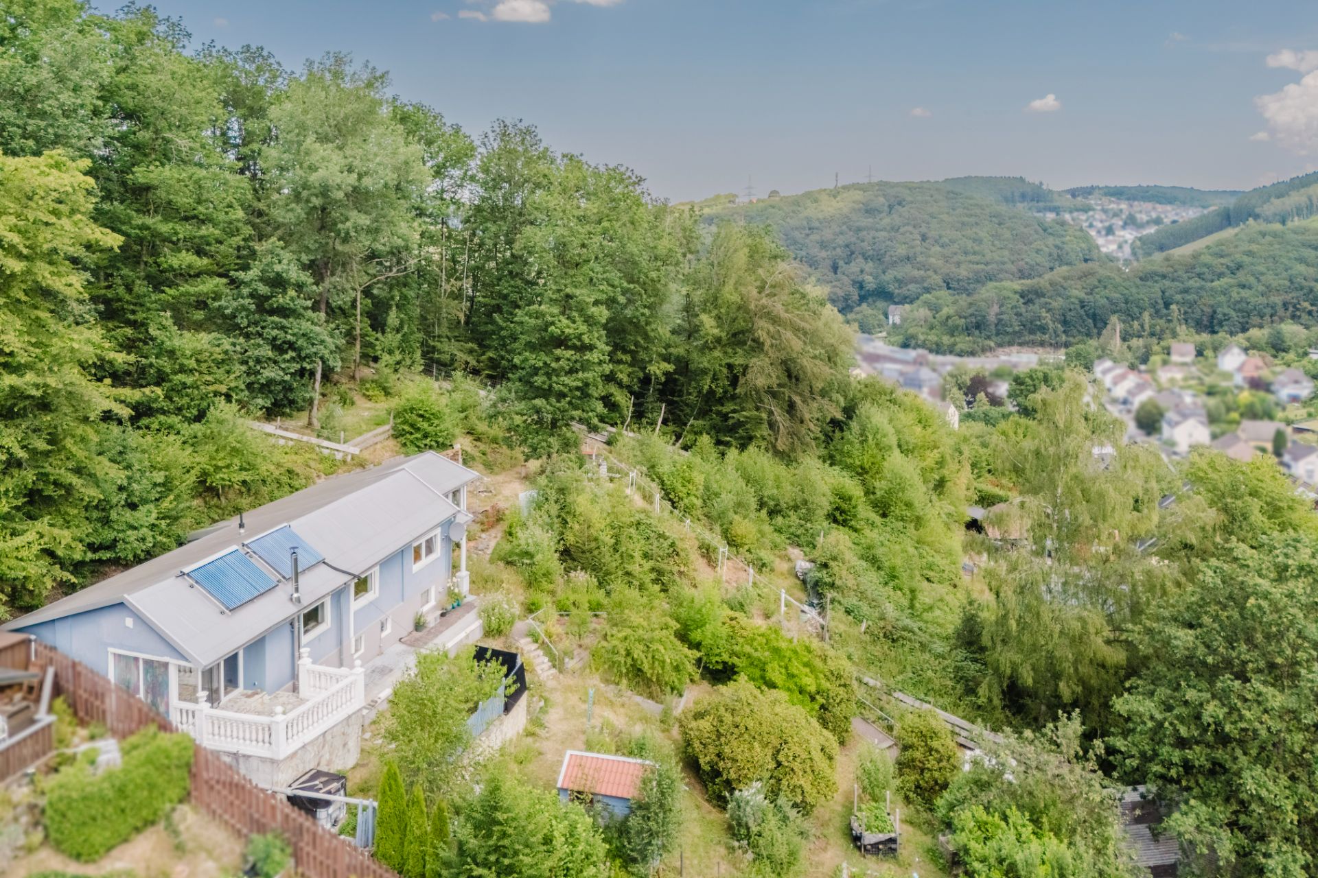 Haus mit weißer Terrasse am Waldrand auf einem bewaldeten Hang, Blick auf Nachbarhäuser im Tal.