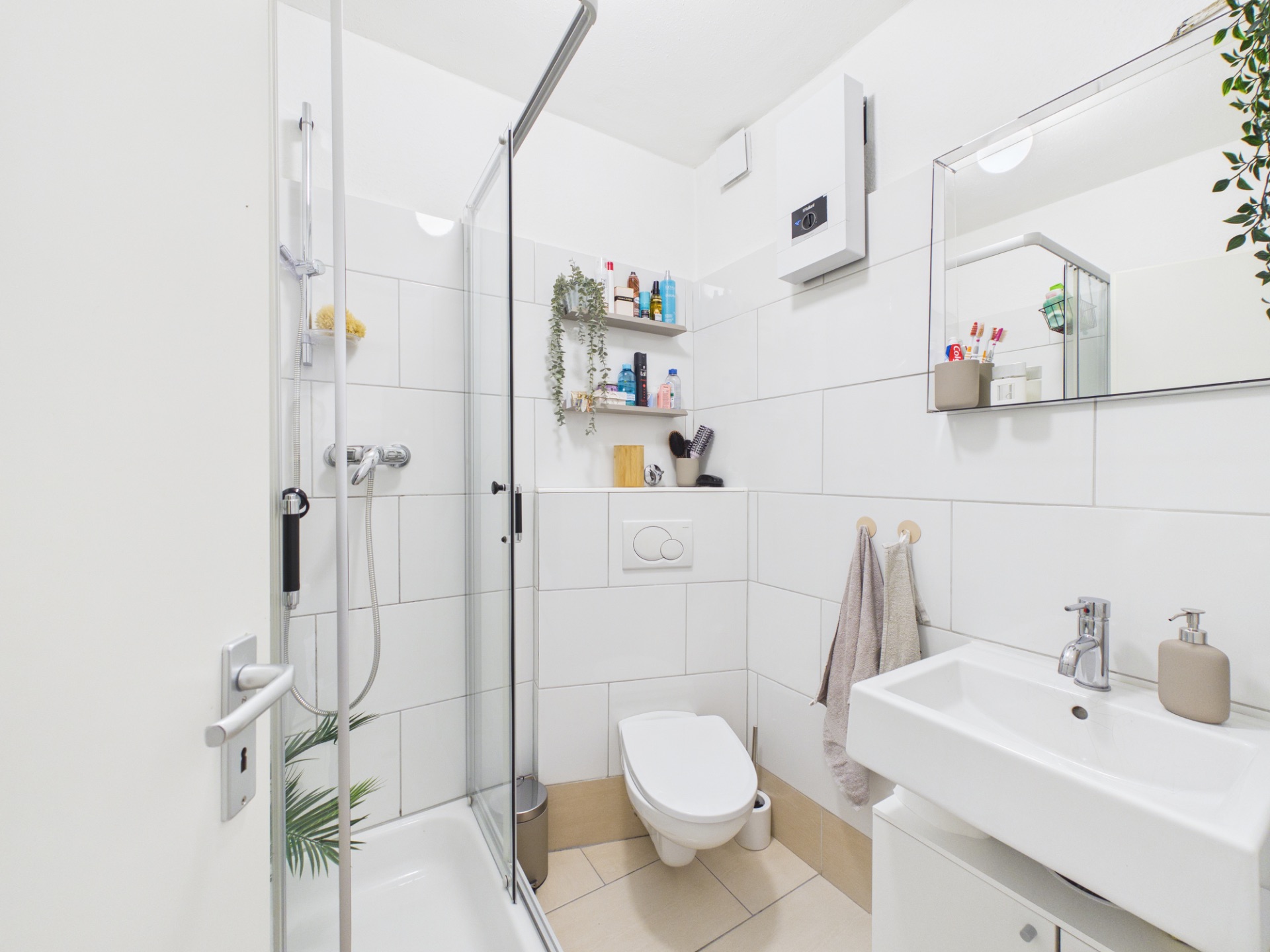 Helles Badezimmer mit Dusche, WC, Waschbecken, Spiegel und Regal mit Pflegeprodukten.