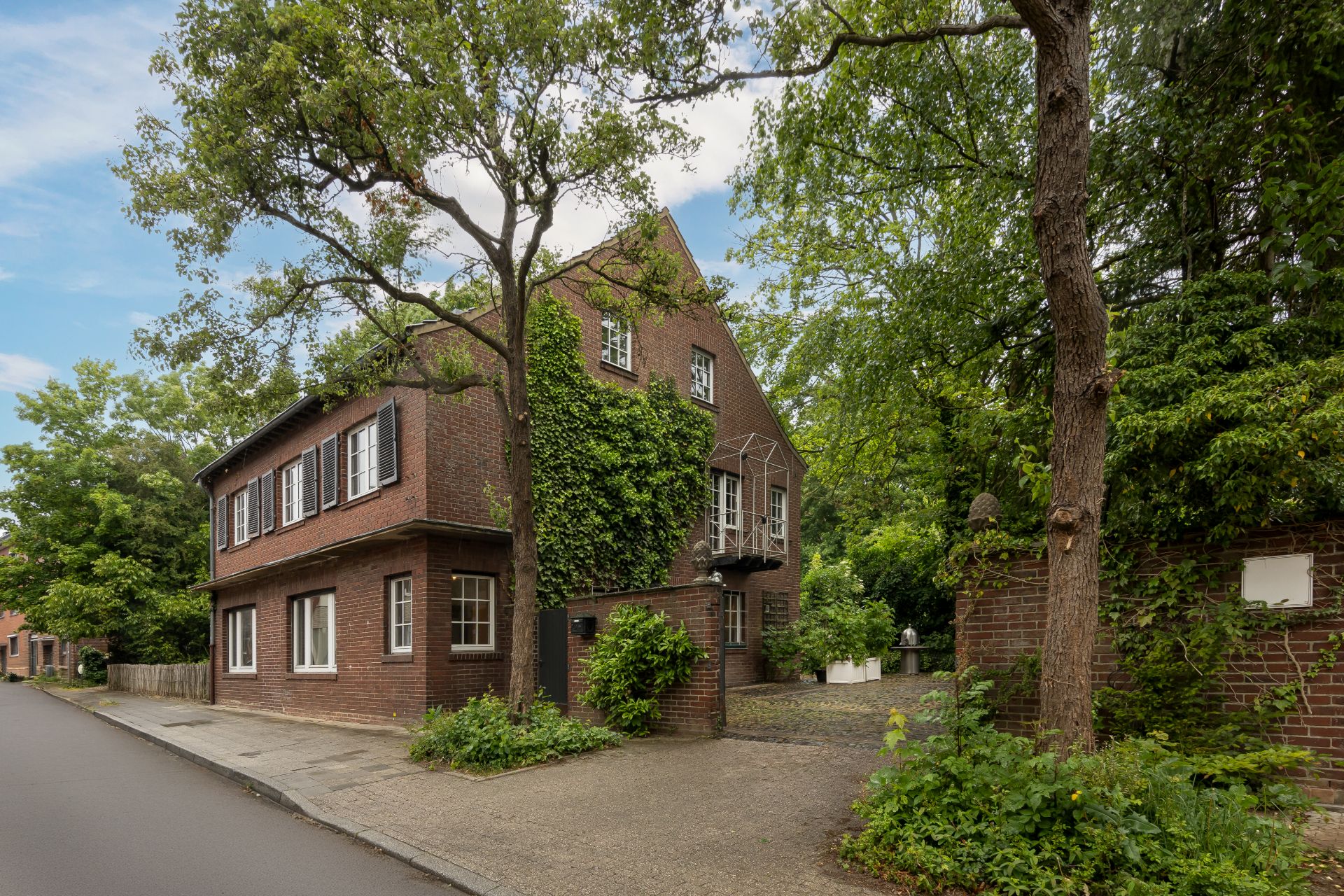 Backsteinwohnhaus mit vielen Fenstern und Kletterpflanzen, umgeben von Bäumen an einer ruhigen Straße.