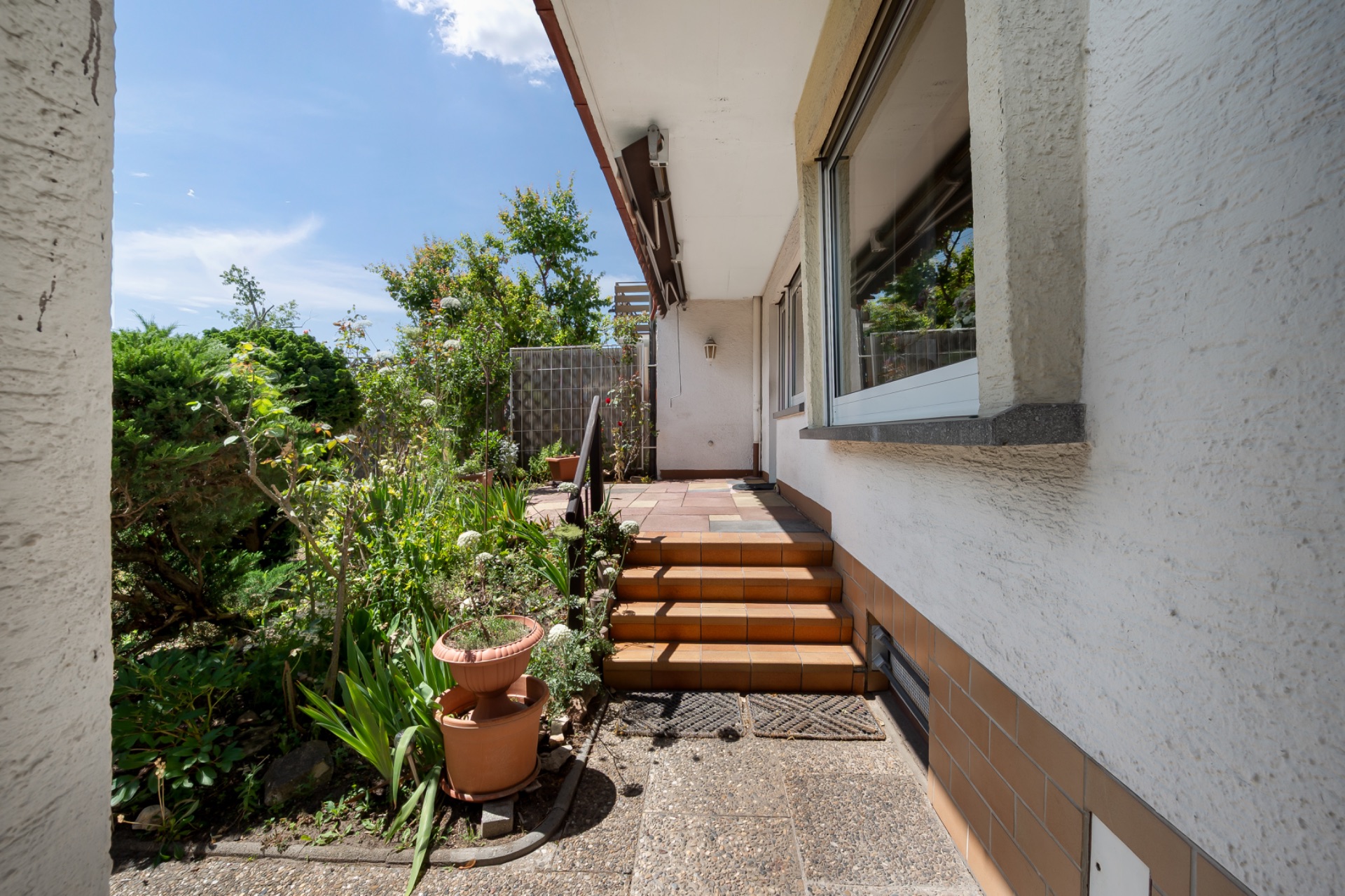 Außentreppe mit Fliesen, Pflanzen und Blick auf einen Garten neben einem Wohnhaus.