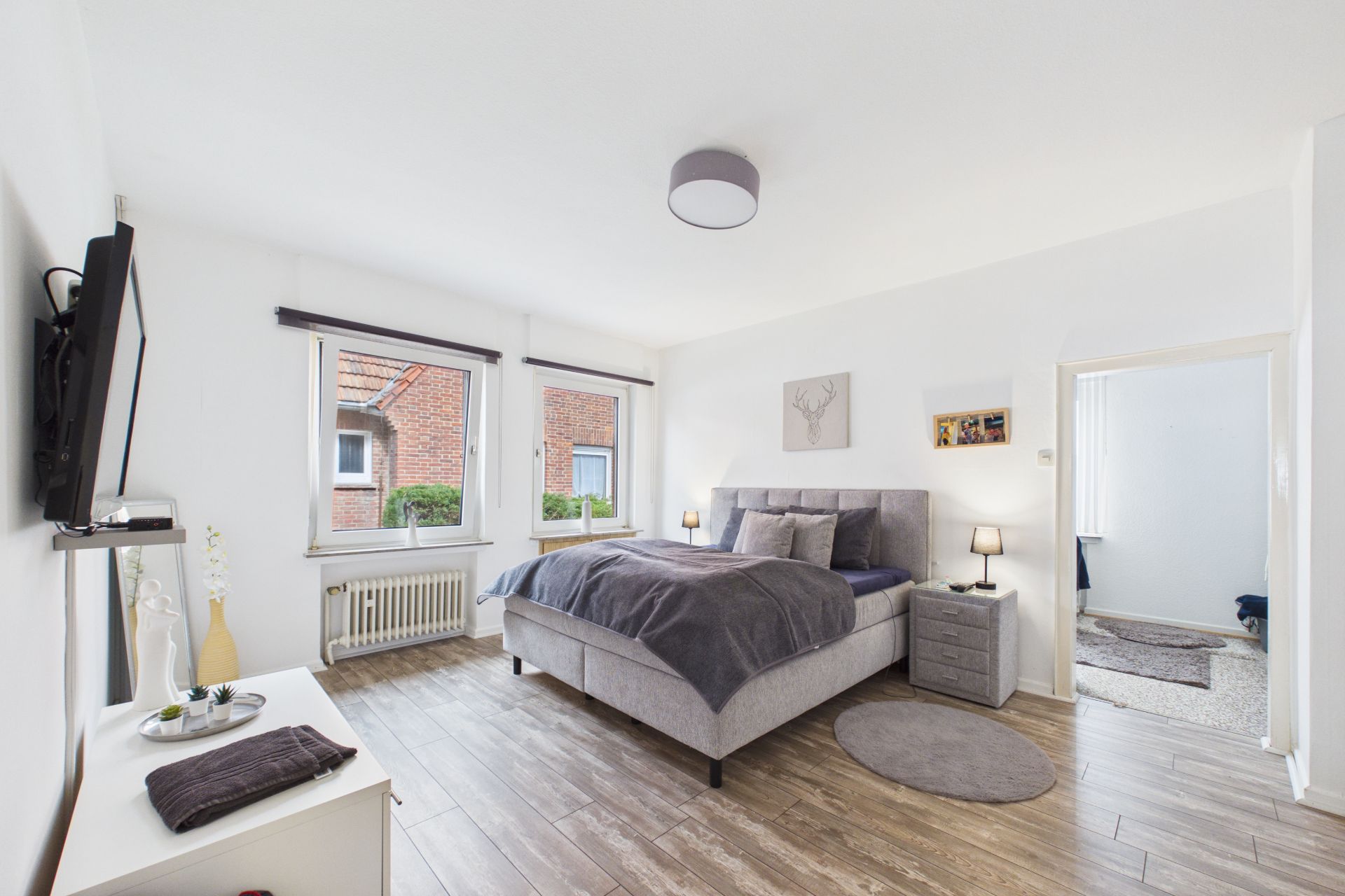 Helles Schlafzimmer mit Doppelbett, zwei Fenstern, Nachttischen und Fernseher an der Wand.
