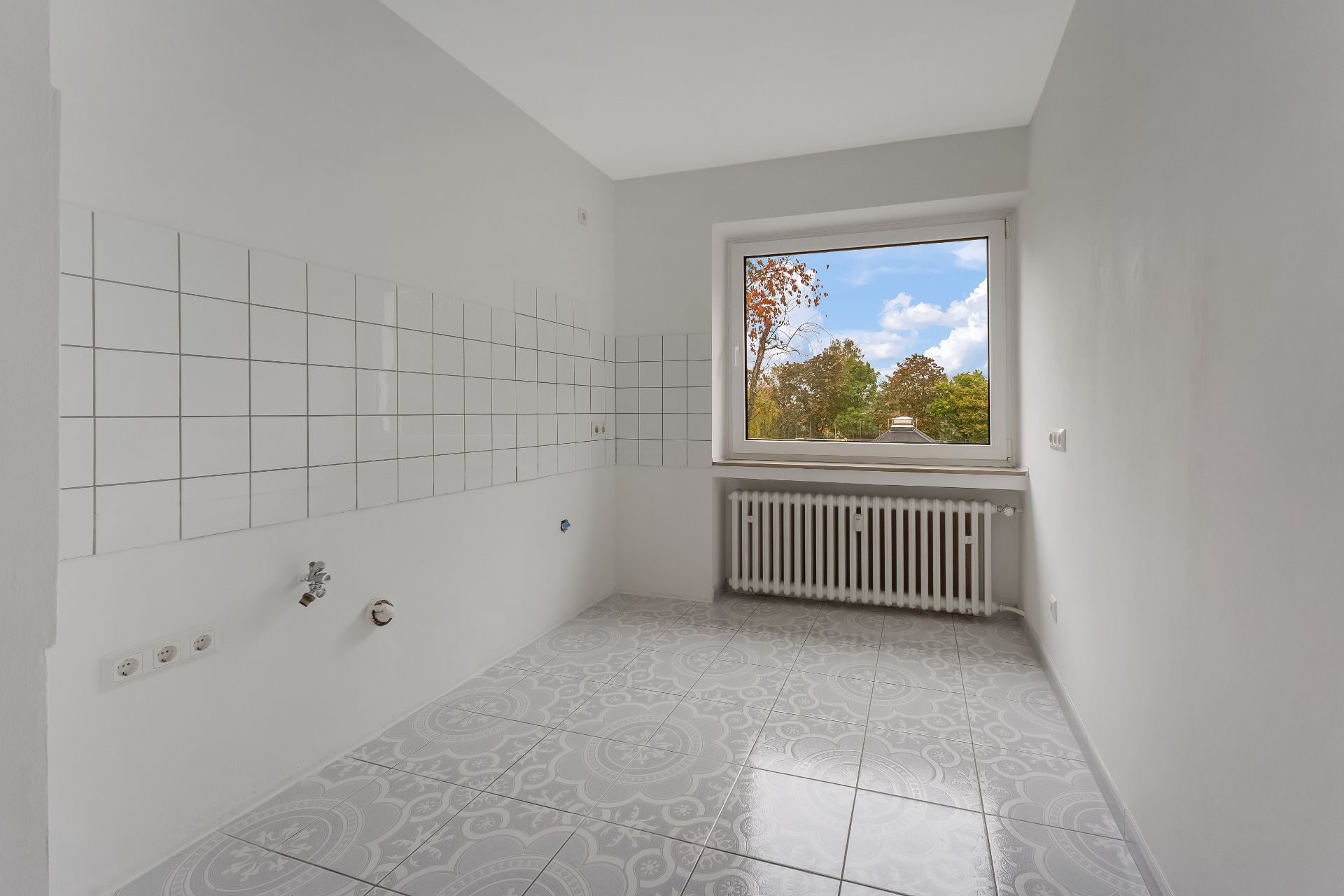 Leerer, heller Raum mit Fliesenboden, weißen Fliesen an der Wand und großem Fenster mit Blick ins Grüne.
