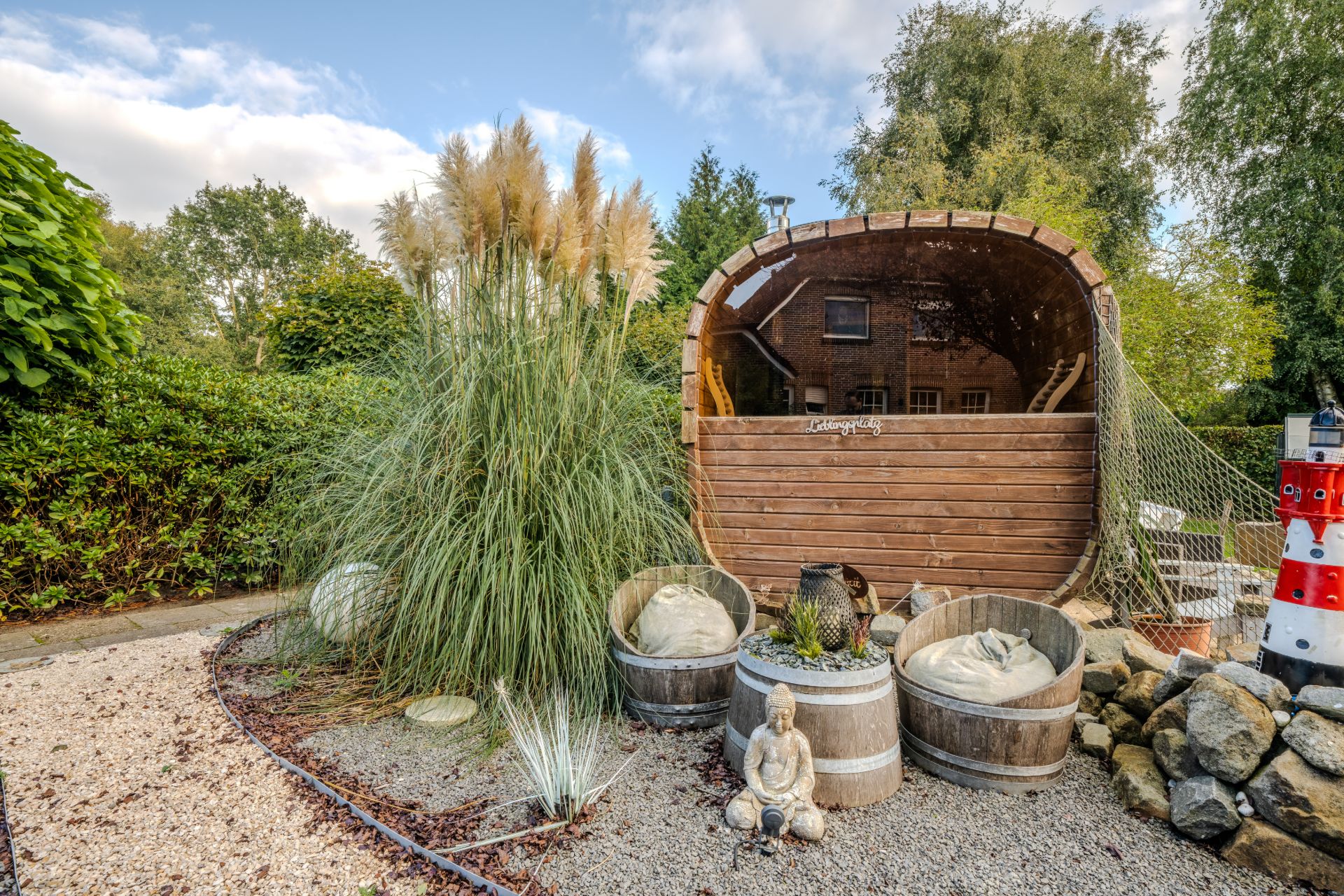 Garten mit Ziergräsern, Holzfass-Sitzmöglichkeit, Dekofässern und Buddha-Figur.