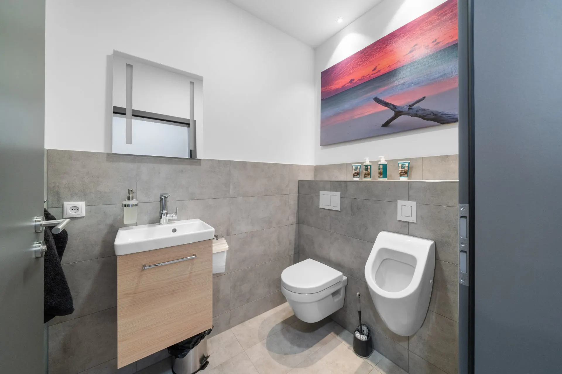 Modernes Badezimmer mit Wandtoilette, Urinal, Waschbecken und Bild an der Wand.